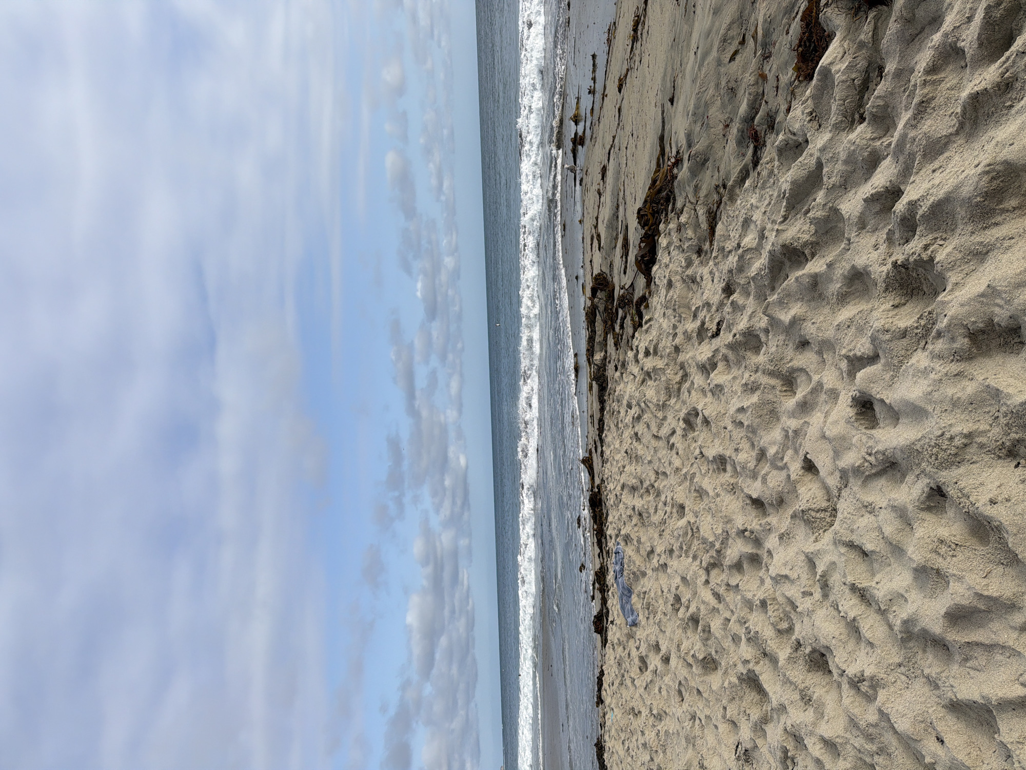 A wide shot captures a serene beach scene in Laguna Beach, California. The foreground is dominated by rippled sand, bearing the marks of footsteps. A blue cloth lies on the sand in the middle ground. Beyond the sand, the ocean stretches out, with waves gently breaking and receding. The horizon is defined by the meeting of the sea and a sky filled with scattered clouds against a pale blue backdrop. The overall atmosphere is tranquil and natural.