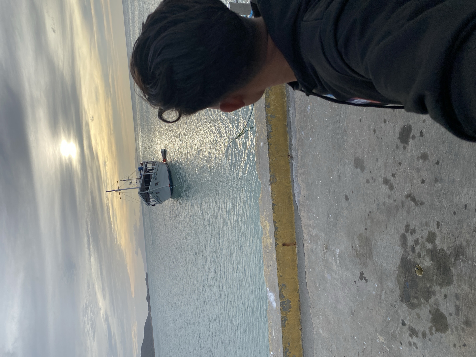 The scene depicts a waterfront area in Juan Griego, Venezuela, under an overcast sky with diffused sunlight. In the foreground, a concrete pier or dock occupies the lower half of the image, exhibiting stains and wear. A yellow-painted concrete curb, running horizontally, separates the pier from the water. A person, viewed from the back and positioned on the right side of the foreground, is wearing a black hooded sweatshirt. This individual has dark, short hair with a distinct curl on the right side and is looking out over the water.
The midground features calm, light blue-green water that reflects the sky. A white, medium-sized boat, possibly named "CONSUELO" with a visible registration number "P-1017" on its bow, is moored in the water to the right of the center. The boat features a cabin, a mast with rigging, and an antenna. A red buoy is visible near its hull, and a small dinghy is tethered to its stern. A green rope extends from the water towards the right side of the concrete pier.
In the background, a line of land masses or hills defines the horizon on the left. The sky is predominantly grey with scattered white clouds, and a bright, yellowish glow from the diffused sun is visible in the upper-right quadrant, indicating either early morning or late afternoon.