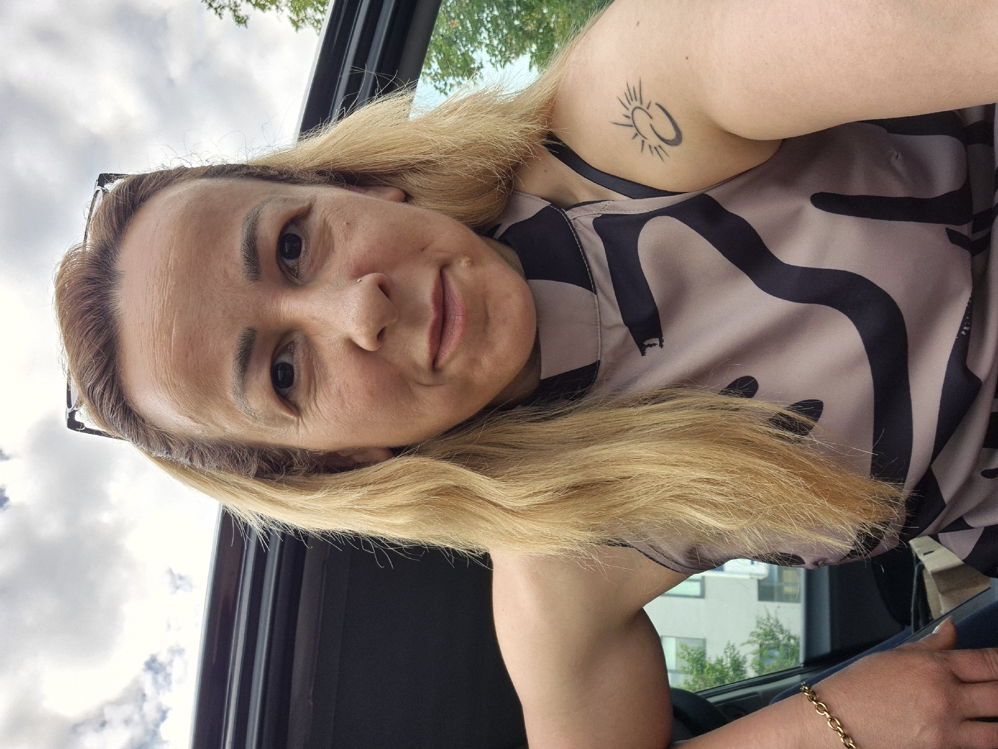 A female subject is captured in a close-up, chest-up self-portrait, positioned at what appears to be a vehicle's open door or window. The subject has long, blonde hair, some strands appearing windblown. Glasses with black frames are perched on top of her head. Her face shows visible wrinkles on the forehead and around the dark eyes, and a small stud is present in her left nostril. A small mole is visible below the right corner of her mouth. She is wearing a sleeveless top featuring a light brown/taupe base with large, irregular black abstract shapes. On her left upper arm, a black ink tattoo depicts a stylized sun with rays emanating from a crescent shape, positioned above a solid crescent moon. A gold-colored chain bracelet is worn on her left wrist. The background consists of a cloudy sky above, with green tree foliage and a light-colored building with dark windows partially visible on the right, indicating an outdoor urban or suburban setting in the city of Unknown, Unknown. The dark interior or frame of a vehicle is visible on the left side of the image.