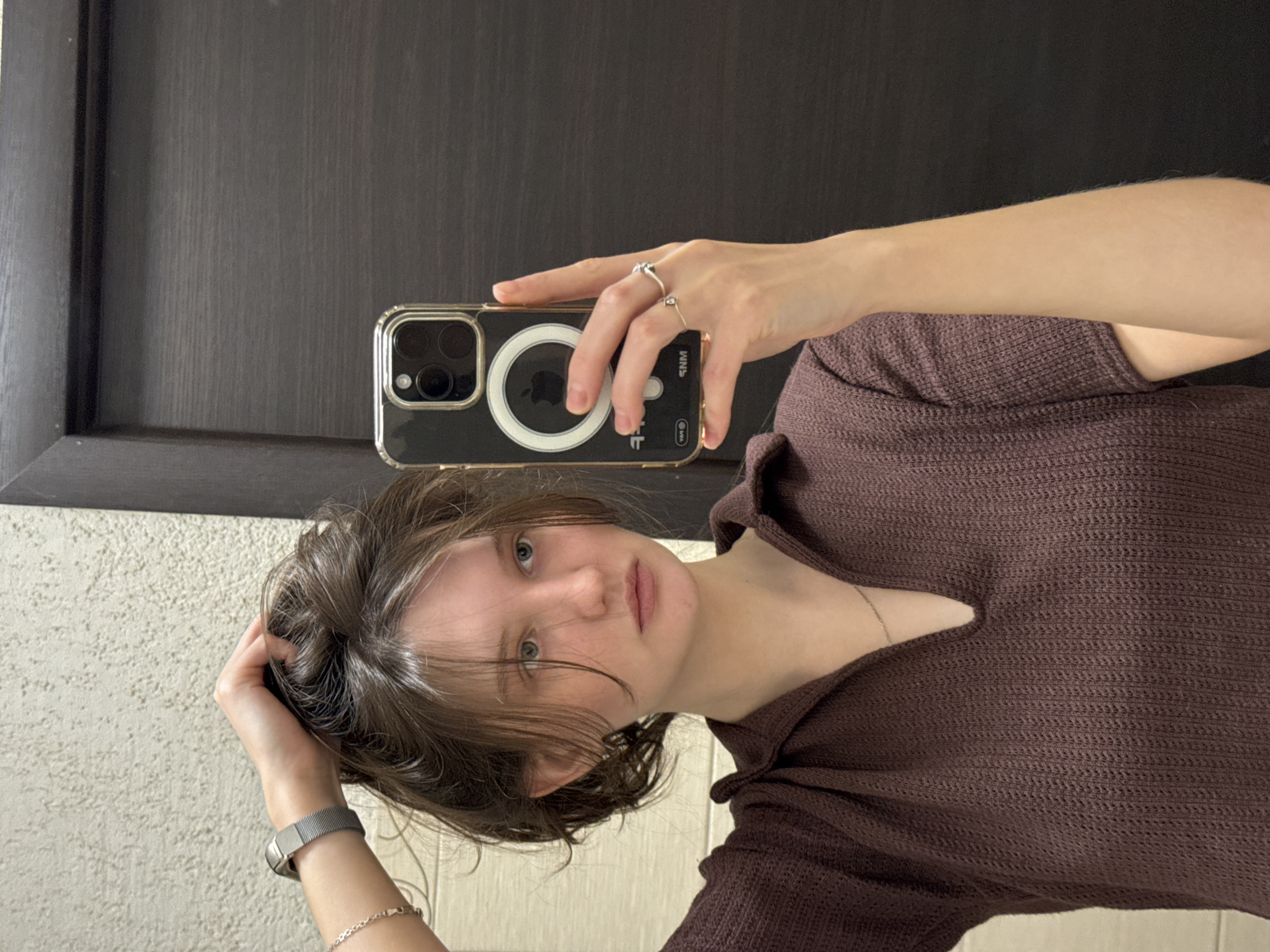 A young woman takes a selfie with her iPhone 14 Pro, clad in a clear case with a magnetic ring, against the backdrop of what appears to be a restroom. She wears a brown knit top with a small V-neck cutout. On her left wrist, she sports a silver watch with a mesh band and a delicate silver bracelet. Her right hand, holding the phone, is adorned with two simple silver rings. Her hair is brown and styled in a casual, slightly disheveled manner. The background features a dark brown wooden door and a light-colored wall with a subtle textured pattern. There is no information available in the image to determine the location as Perm, Russia.