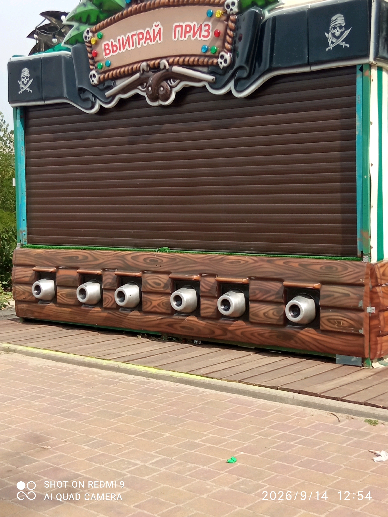 The image depicts a game booth with a pirate theme located in Rostov-on-Don, Russia. The booth features a brown roller-shutter door at the back, with a decorative wooden facade in the foreground. This facade has seven openings, each containing a silver-colored barrel or cannon-like object. Above the shutter, a sign with red Cyrillic text that reads "ВЫИГРАЙ ПРИЗ" (Win a Prize) is visible, adorned with pirate skull and crossbones motifs and colorful lights. The booth is situated outdoors, with a paved walkway in the foreground and wooden decking behind the booth. The overall setting suggests an amusement park or a public recreational area.