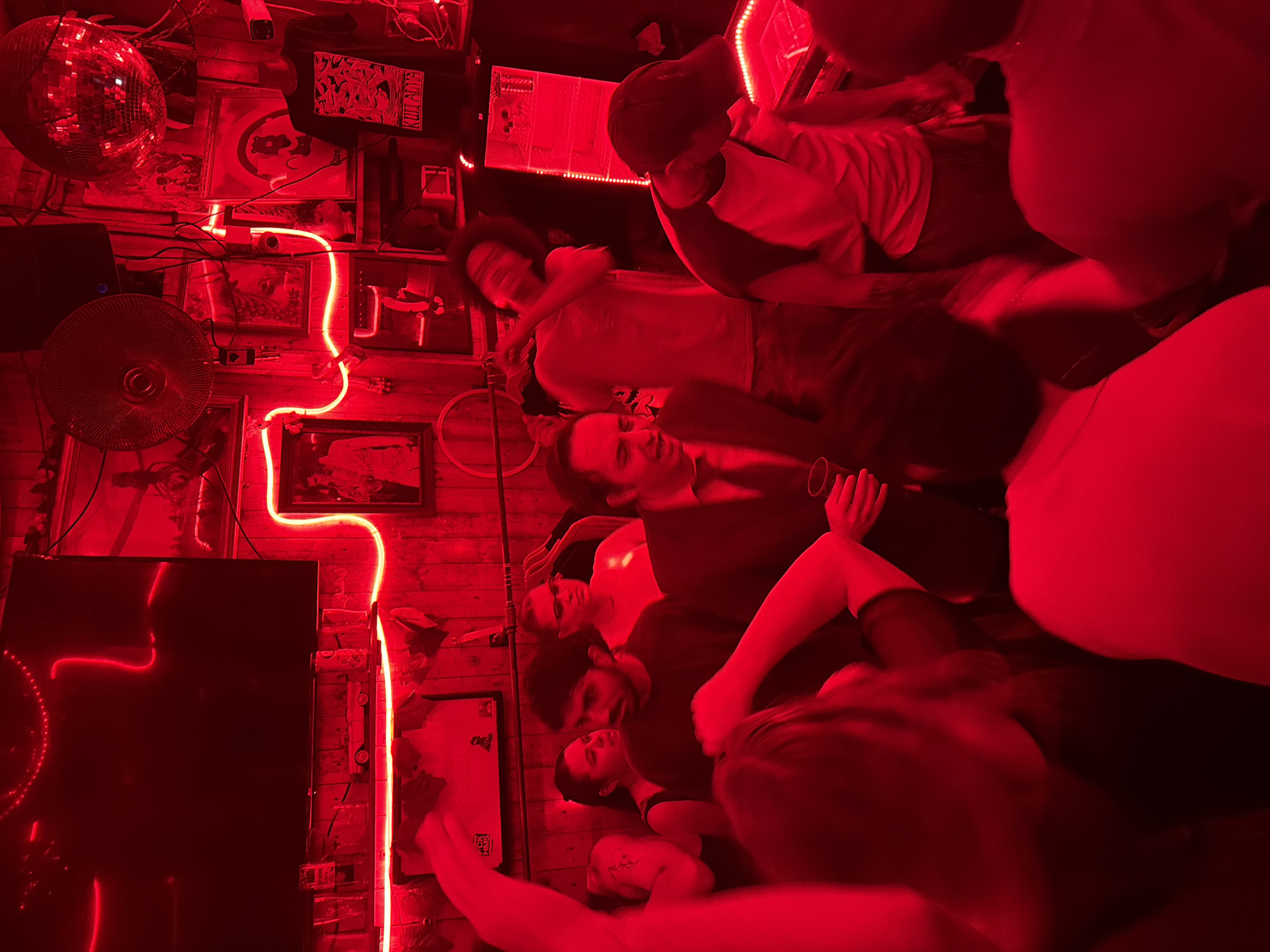 This image captures a crowded indoor establishment, identified as being in Tel Aviv, Israel. The scene is enveloped in a strong, pervasive red light, originating from extensive neon tubing and strip lights that outline architectural features and adorn the walls.
The environment consists of wooden plank walls densely covered with framed pictures and posters, suggesting a themed bar or club. A large, dark flat-screen television is mounted on the upper left, beneath which a shelf displays a miniature vintage car and several drinking glasses. In the upper right section, a large disco ball hangs, alongside a wall-mounted circular fan and various framed artworks. Neon lights form distinct patterns, including a vertical "2" or "S" shape and horizontal lines across the walls. A dark t-shirt, possibly on a mannequin or hanger, is visible on the right with the text "KILL SOME" printed on it, positioned near an illuminated vertical display unit. A partially visible sign reads "F**K HMS" in the mid-ground.
Approximately 10-15 individuals are present, creating a dense social gathering. In the central foreground, a man wearing a dark suit jacket and tie holds a clear glass, smiling and looking towards his left. Behind him, a man with dark, curly hair in a light t-shirt is depicted with motion blur, suggesting movement or dancing, his right arm raised across his chest. To the far right, a man in a dark baseball cap and a white-and-dark raglan shirt is bent over, seemingly interacting with an illuminated, red-lit surface, possibly a pinball machine or arcade game. In the mid-ground, a bearded man in a dark t-shirt looks intently to his right, while behind him, two women are partially visible, one wearing glasses. Numerous other individuals are present in the foreground and background as obscured or partial figures, collectively indicating an active social scene involving conversation, drinking, and movement.