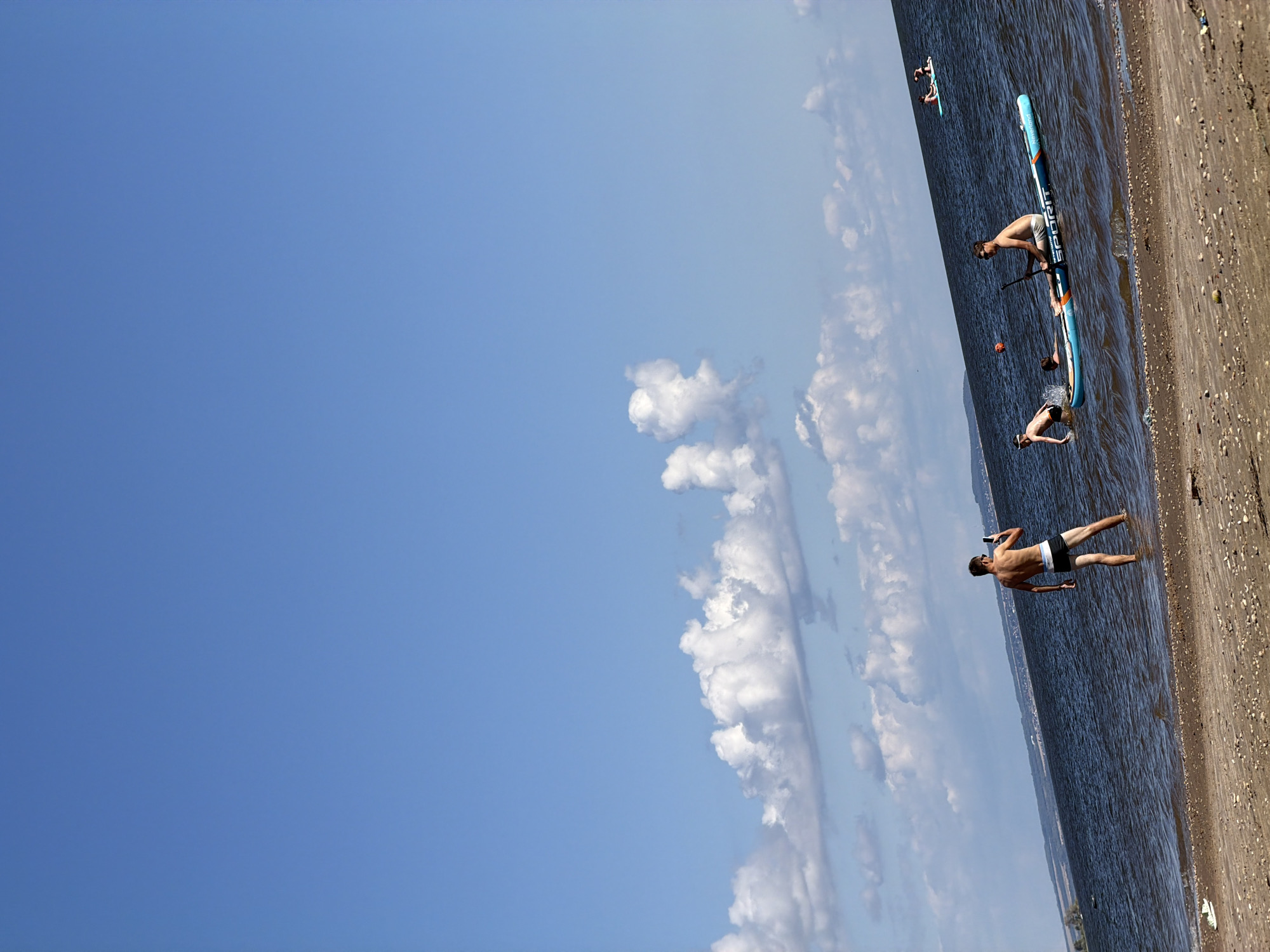 The image depicts a serene waterscape on a bright, sunny day. The sky is a vibrant blue, adorned with scattered, fluffy white clouds. The water, a deep blue, gently laps at a sandy shore littered with pebbles and debris.
In the foreground, a lone individual stands near the water's edge, facing away from the camera and holding a mobile phone, seemingly taking a picture. The person is wearing dark swim trunks.
Further out in the water, a stand-up paddleboard with the text "Neverland Spirit" is visible. On this board, two individuals are present: one is seated, holding a paddle, while another appears to be in a prone position, possibly falling or attempting to get up. A third person is in the water next to the paddleboard.
In the distance, on another paddleboard, two more individuals are visible, enjoying the water. The shoreline in the far distance is marked by a thin line of vegetation and some structures, suggesting a developed area beyond the natural landscape. The overall atmosphere is one of leisure and outdoor recreation on a clear, summer day.