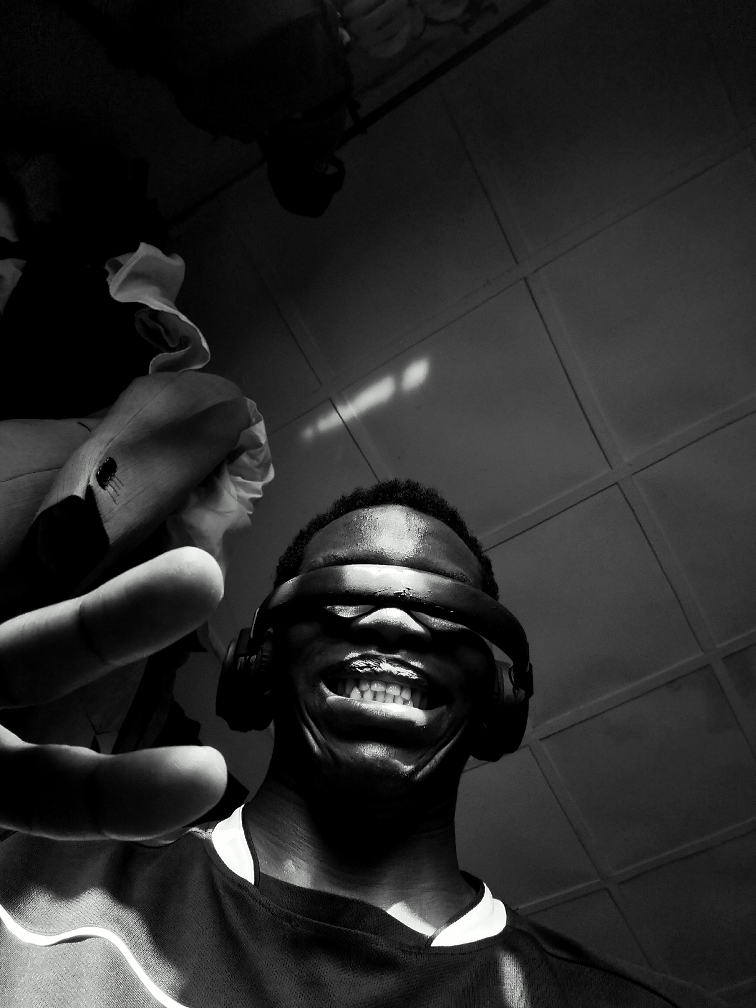 A low-angle, black-and-white shot of a dark-skinned person wearing headphones that are pulled over the eyes like a blindfold. The person is smiling broadly, revealing their teeth. Their left hand is outstretched toward the viewer. The person is wearing a dark, collared shirt. Above the person, the background is a patterned surface, possibly a tiled ceiling or wall. The lighting is dramatic, with a bright streak of light on the tiled surface and parts of the hand and forehead. The location of Keffi, Nigeria is not visually discernible from the image.
