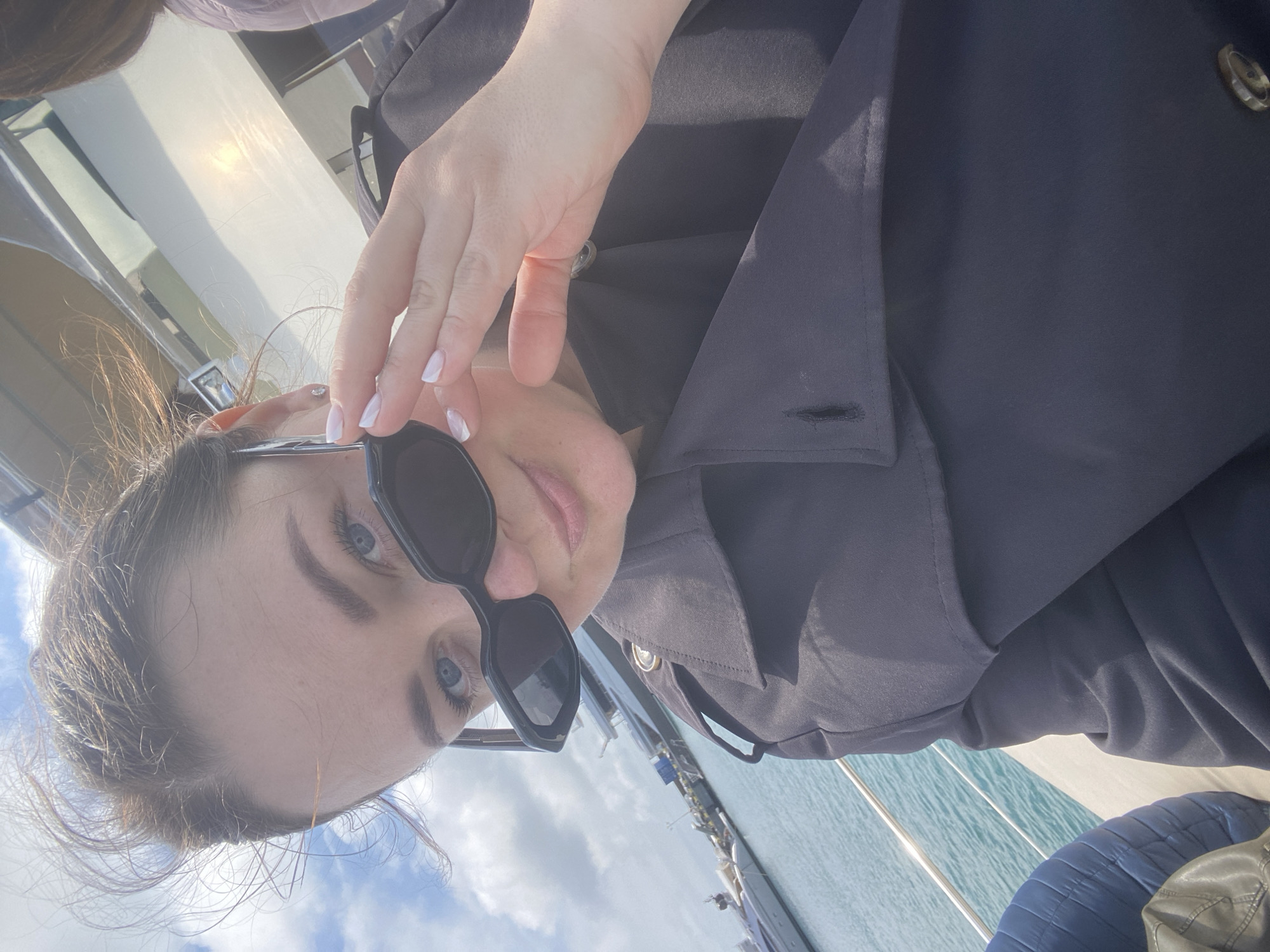 A woman with blue eyes and fair skin is holding up her sunglasses with her left hand. She is wearing a dark-colored coat. The background shows a body of water and what appears to be a yacht or boat. Given the location, it is reasonable to assume the body of water is part of the Black Sea. The sky is visible with some clouds.