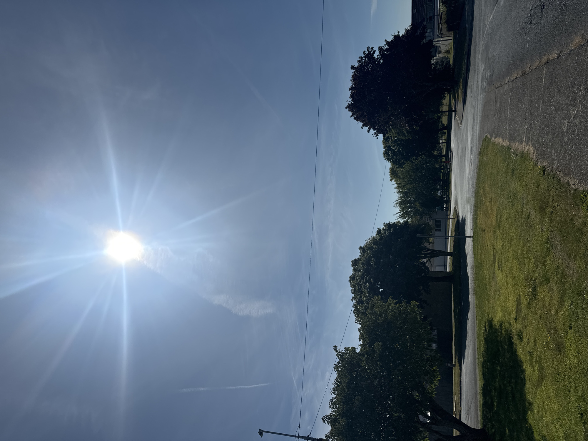 The image captures a bright, sunny day with the sun positioned high in a clear blue sky, creating lens flare and radiating beams. A contrail is visible in the upper left quadrant of the sky. In the foreground, a paved walkway curves to the right, bordered by a patch of green grass. Beyond the grass, a paved road winds through a neighborhood lined with trees. Several mature trees with lush green foliage are scattered throughout the scene, casting shadows on the road and grass. A street lamp stands on the left side of the image. In the background, houses and buildings are visible amongst the trees. The overall scene depicts a suburban or residential area in Waterford, Ireland, on a clear day.
