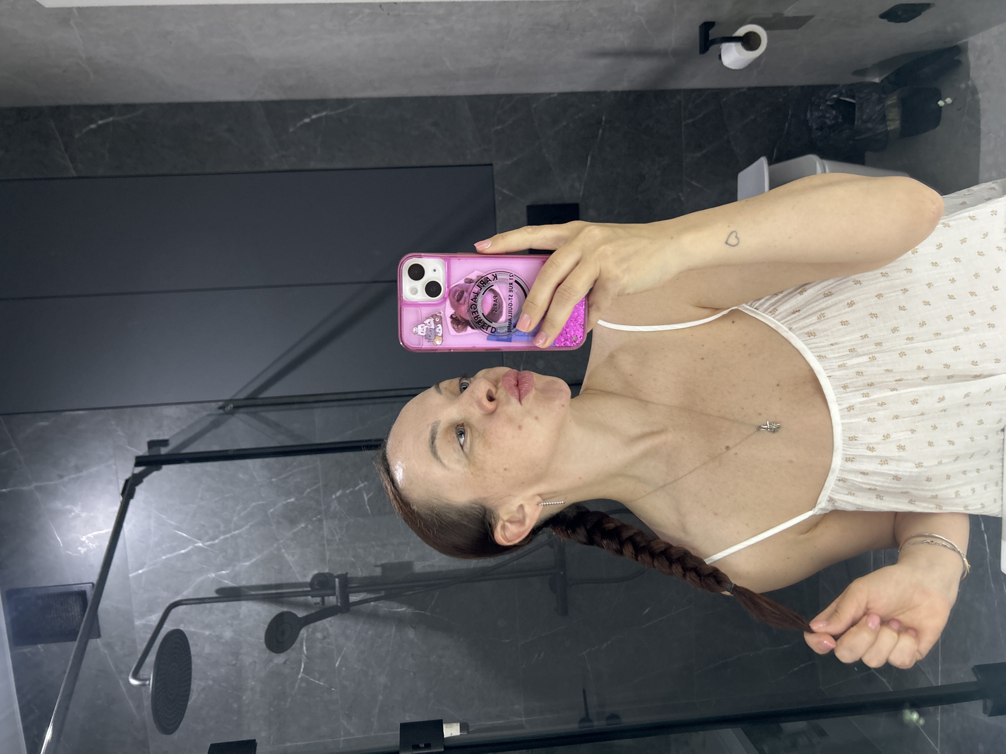 A candid mirror selfie captures a female in a modern bathroom, reportedly located in Yekaterinburg, Russia. The individual, who has freckles and dark brown hair styled in a single braid extending over her left shoulder, is wearing a white camisole with thin straps and a subtle patterned texture. She is accessorized with small silver stud earrings, a delicate silver necklace featuring an intricate pendant, and two thin silver bracelets on her left wrist. A small outline of a heart is tattooed on her right forearm. She holds a pink smartphone with a decorative case featuring glitter, a teddy bear, and text (reflected to show 'PARIS' and '24 RUE ST-GUILLAUME'), positioning it to take the photo while making a "kissy" face towards her reflection. Her left hand gently holds the end of her braid. The bathroom environment features dark grey marble-effect wall tiles with white veining and dark matte wall panels. Visible elements include a glass shower enclosure with dark framing, a black rain shower head, a handheld shower, a wall-mounted soap dispenser, and a square ventilation grate. To the right, a black toilet paper holder with a roll of white toilet paper is visible, alongside a small white waste bin partially obscured by the subject's arm.
