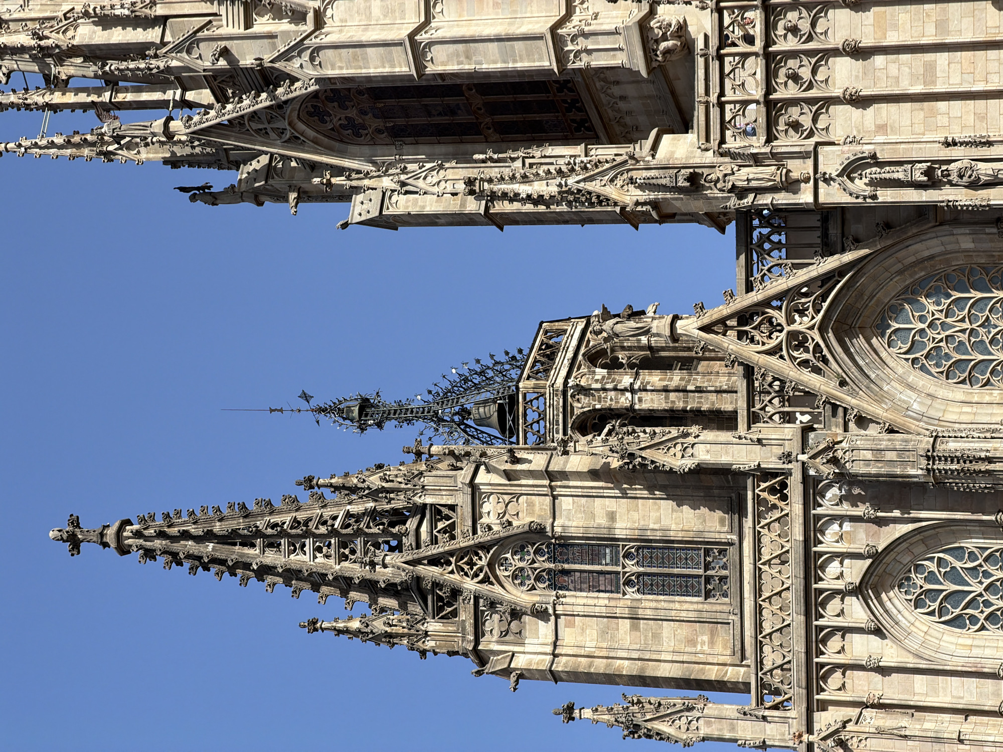 The image shows a close-up, low-angle view of the spires and facades of the Barcelona Cathedral, also known as the Cathedral of the Holy Cross and Saint Eulalia, located in the Gothic Quarter of Barcelona, Spain. The architecture is Gothic, characterized by intricate stone carvings, pointed arches, spires, and ornate decorative elements. Several spires, some topped with crosses, rise towards a clear blue sky. Detail is visible in the stonework, including gargoyles, statuary, and tracery within the windows. The stone appears weathered, suggesting the age of the structure. Sunlight casts shadows on the facade, highlighting the three-dimensional details of the architecture. No people are clearly visible in the image. The environment is dominated by the grand stone edifice and the bright sky.