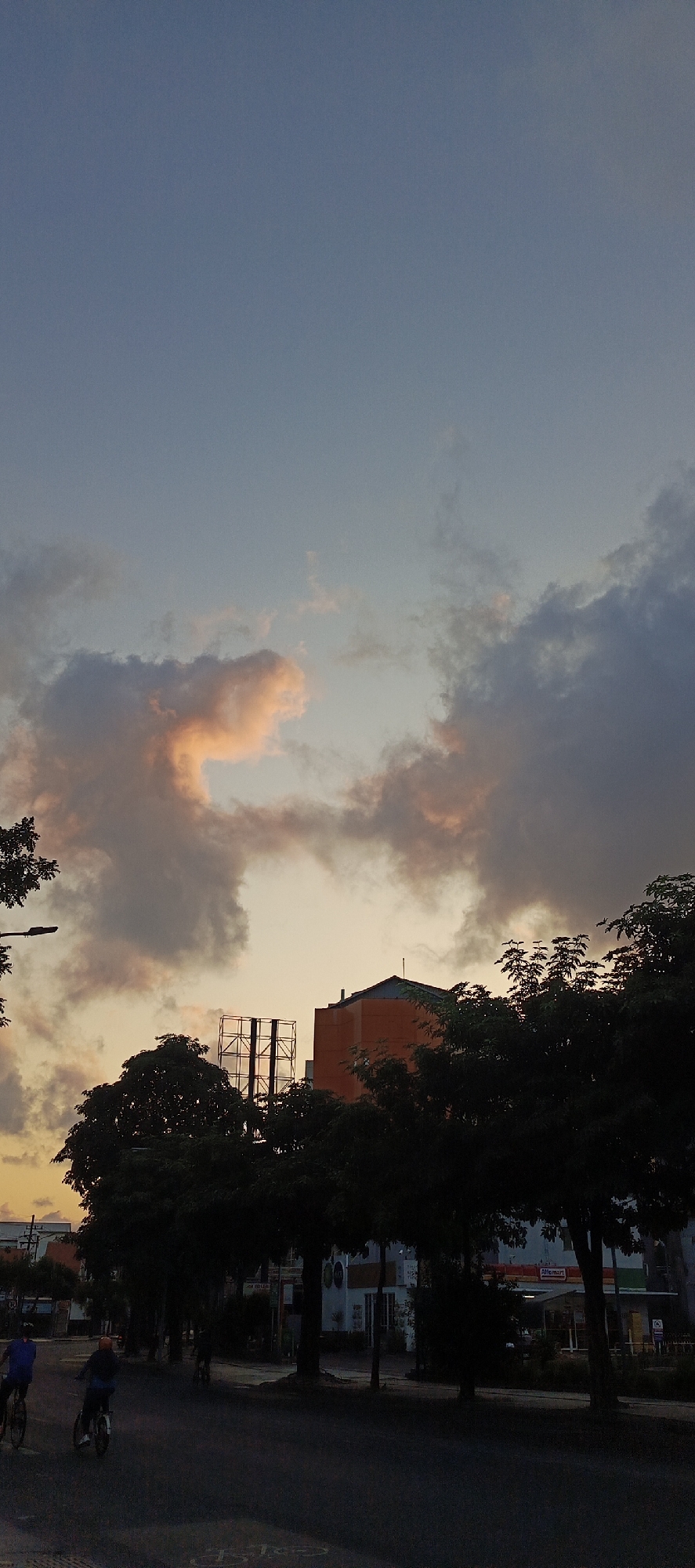 The image captures a street scene in Surabaya, Indonesia, during what appears to be sunset. The sky is a gradient of soft blue to pale orange, with dramatic clouds tinged with pink and gray. Silhouetted against the sky are trees and buildings. In the foreground, a paved street is visible with two individuals cycling. One cyclist, wearing a blue shirt, is on the left, and another, in a blue jacket, is to their right. Further down the street, other figures and vehicles are faintly discernible. A dark-roofted building with orange accents and a large metal structure, possibly a billboard frame, are prominent in the mid-ground, partially obscured by trees. The overall atmosphere is tranquil, characteristic of an urban evening in Surabaya.
