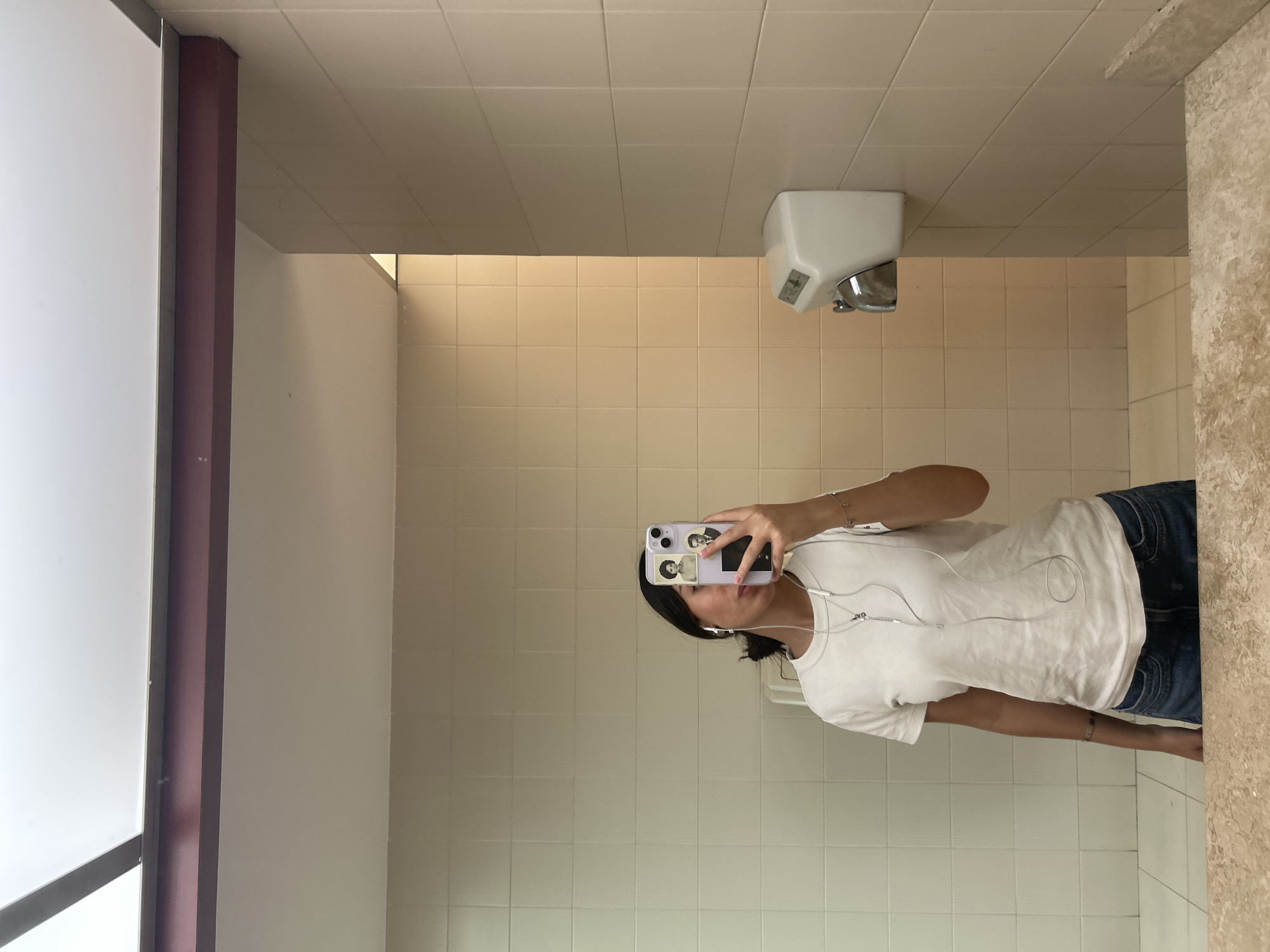 An individual is captured in a mirror selfie within an indoor setting, likely a public restroom or utility area, characterized by tiled walls and ceiling. The image is oriented sideways, with the person standing upright relative to the mirror, holding a light purple smartphone with both hands to take the picture.
The person is visible from the upper thighs upwards, wearing a white short-sleeved t-shirt and blue jeans. Dark hair is visible, pulled back. Wired white earbuds are plugged into the phone, with the wires draped over the person's chest. A bracelet is worn on the right wrist. The back of the smartphone case features two small, vintage-style black and white portrait stickers.
The background reflected in the mirror shows light beige/cream square tiles covering the wall behind the person. Above the tiled wall is a light-colored, possibly white, ceiling with square ceiling tiles. A white, wall-mounted hand dryer with a metallic spout is visible in the upper right portion of the background, attached to the tiled wall. A small, greenish label on the hand dryer has illegible text. To the left of the reflected scene, a vertical dark red column or partition is visible next to a large white panel. On the far right, a textured, light-brown surface forms the edge of the mirror or an adjacent wall. The lighting appears to be even and artificial.