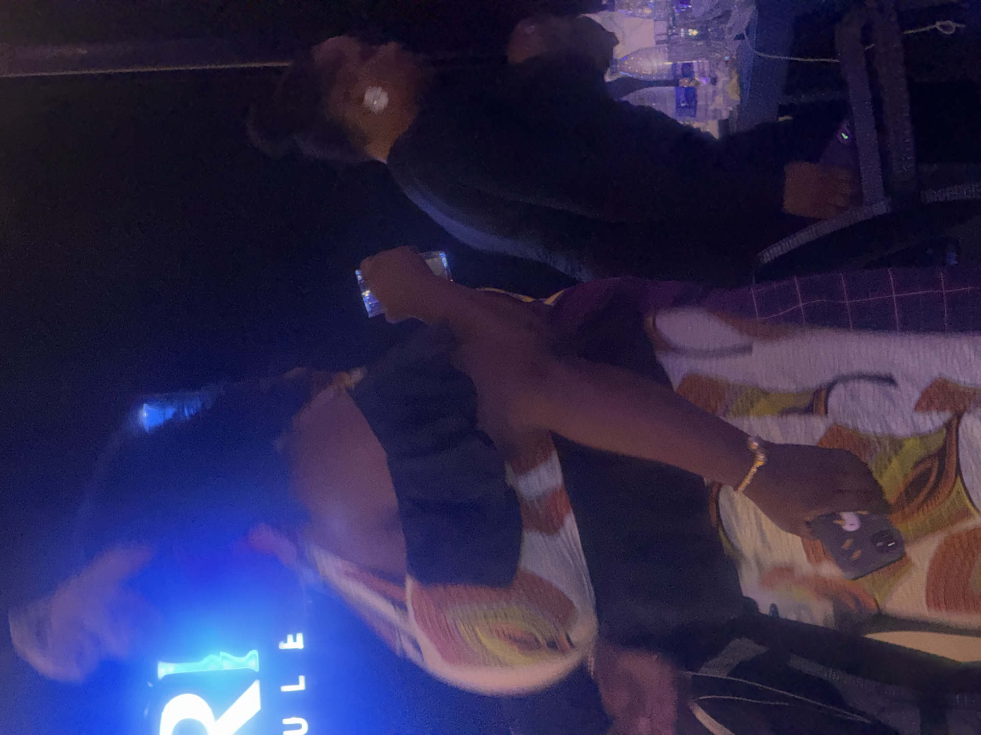 The image depicts a dimly lit indoor scene, possibly a club or event space, in Abuja, Nigeria. The focus is on two individuals, with the primary subject being a woman in the foreground wearing an off-the-shoulder top with a vibrant, abstract pattern in shades of orange, yellow, and white. She is holding a smartphone in her right hand, which is adorned with a gold bracelet. Her left arm is raised, obscuring parts of her face and hair. Behind her, another person is visible wearing a black long-sleeved top, with their face turned away and hair in a bun. In the background, a table is laden with bottles of water and possibly other beverages, illuminated by the ambient lighting. The overall atmosphere suggests a social gathering or entertainment venue.