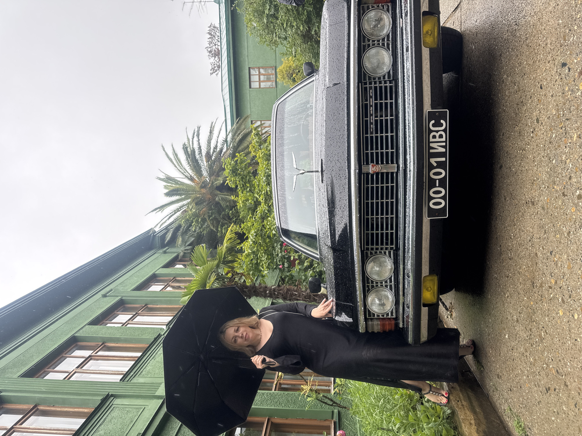A woman stands to the left of a black vintage car, holding a black umbrella. She is wearing a long, black dress and sandals. The car has a chrome grille, round headlights, and a license plate that reads "00-01 ИВС". In the background, a green building with wooden-framed windows is visible, along with palm trees and lush greenery. The scene appears to be in Sochi, Russia, given the architecture and foliage characteristic of the city. The ground is wet, suggesting it is raining. The woman is looking at the camera and appears to be posing with the car.