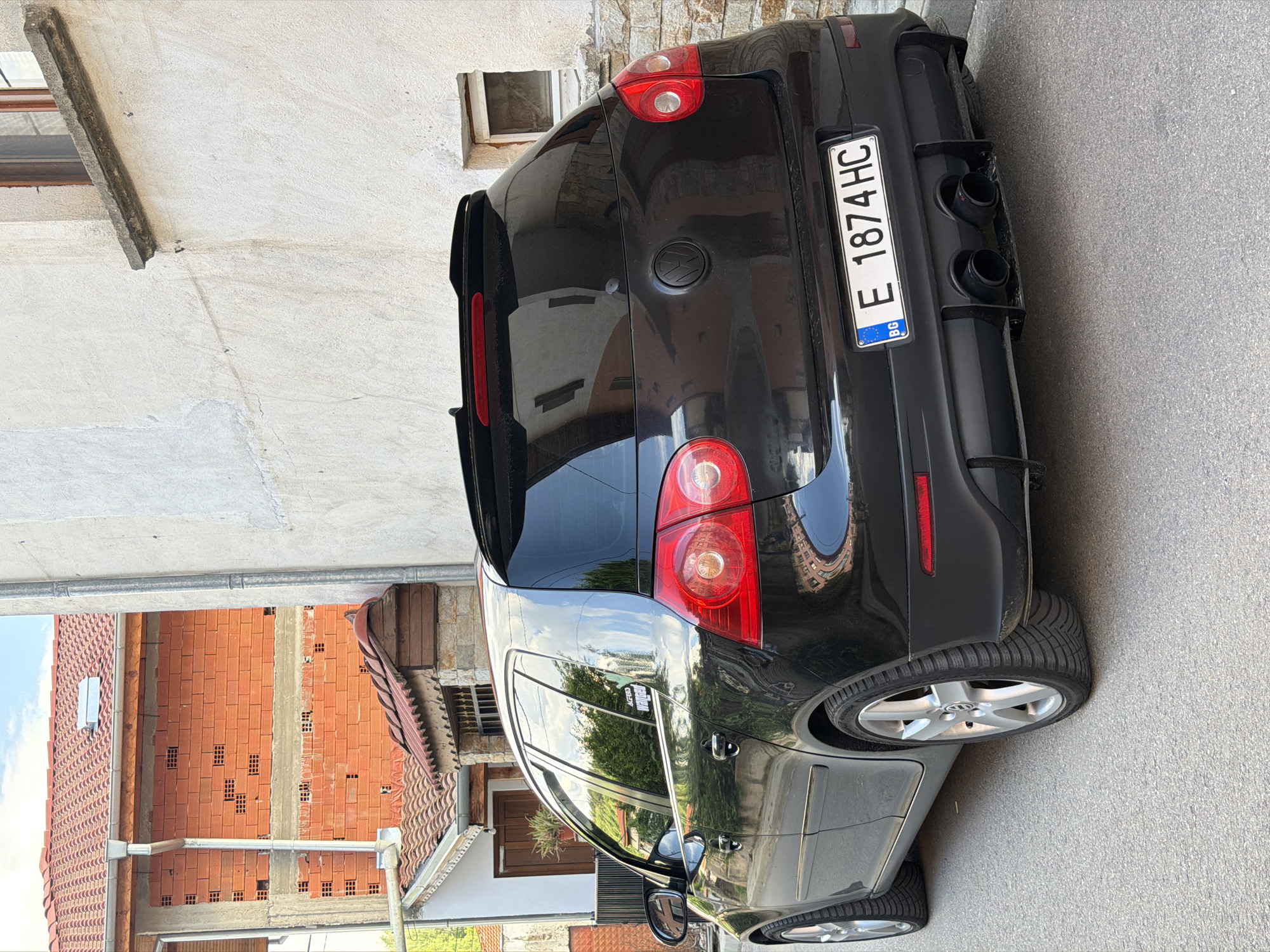 The image displays the rear and right side of a black Volkswagen hatchback, likely a Golf, parked on a paved street. The vehicle is oriented with its rear facing the viewer, angled slightly towards the right. It features dark tinted windows, prominent red taillights, and a black rear diffuser with two large, circular exhaust tips, suggesting a performance model or aftermarket modifications.
The Bulgarian license plate "E 1874 HC" with the "BG" country code is clearly visible on the rear. The car is equipped with silver multi-spoke alloy wheels. Reflections on its glossy black surface and windows show parts of the surrounding environment, including buildings, trees, and a clear blue sky, indicating bright daylight and sunny weather. A small white sticker bearing the text "ACERBIS" and partially "ACTION SPORT" is affixed to the rear passenger side window.
The car is parked alongside a building with a light-colored, possibly stucco, exterior wall that features small, recessed square windows. In the background, other residential buildings with orange-red tiled roofs and exposed red brick walls are visible, some appearing to be under construction or renovation. Gutters and drainpipes are also discernible on these structures. The setting suggests a quiet, residential area in Kochan, Bulgaria. No specific activity or people are depicted; the scene focuses on the parked vehicle within its urban environment.