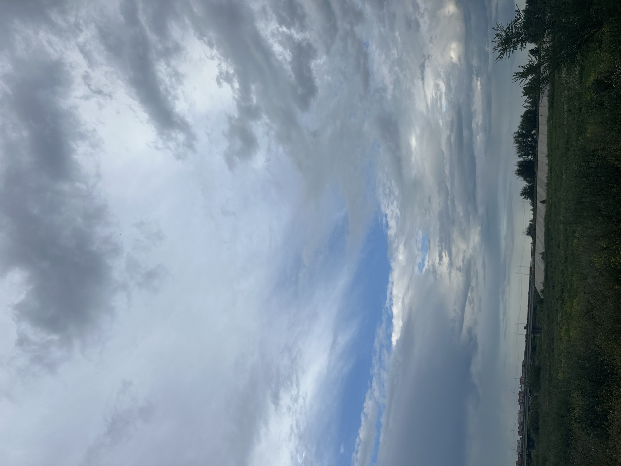 The image captures a dramatic sky with a mix of heavy, dark clouds and lighter, wispy ones, interspersed with patches of bright blue sky. The clouds appear to be moving, suggesting a dynamic weather system. The scene is set outdoors, with a glimpse of a grassy field and trees on the right side of the frame. A low wall or embankment runs horizontally across the middle ground, and beyond that, a few buildings and streetlights are visible in the distance. The time of day is unclear, but the lighting suggests it could be late afternoon or early evening. The overall mood is somewhat somber due to the dark clouds, but the presence of blue sky offers a hint of hope. No people or text are visible in the image. The location is identified as Заречный ауылдық округі, Kazakhstan.
