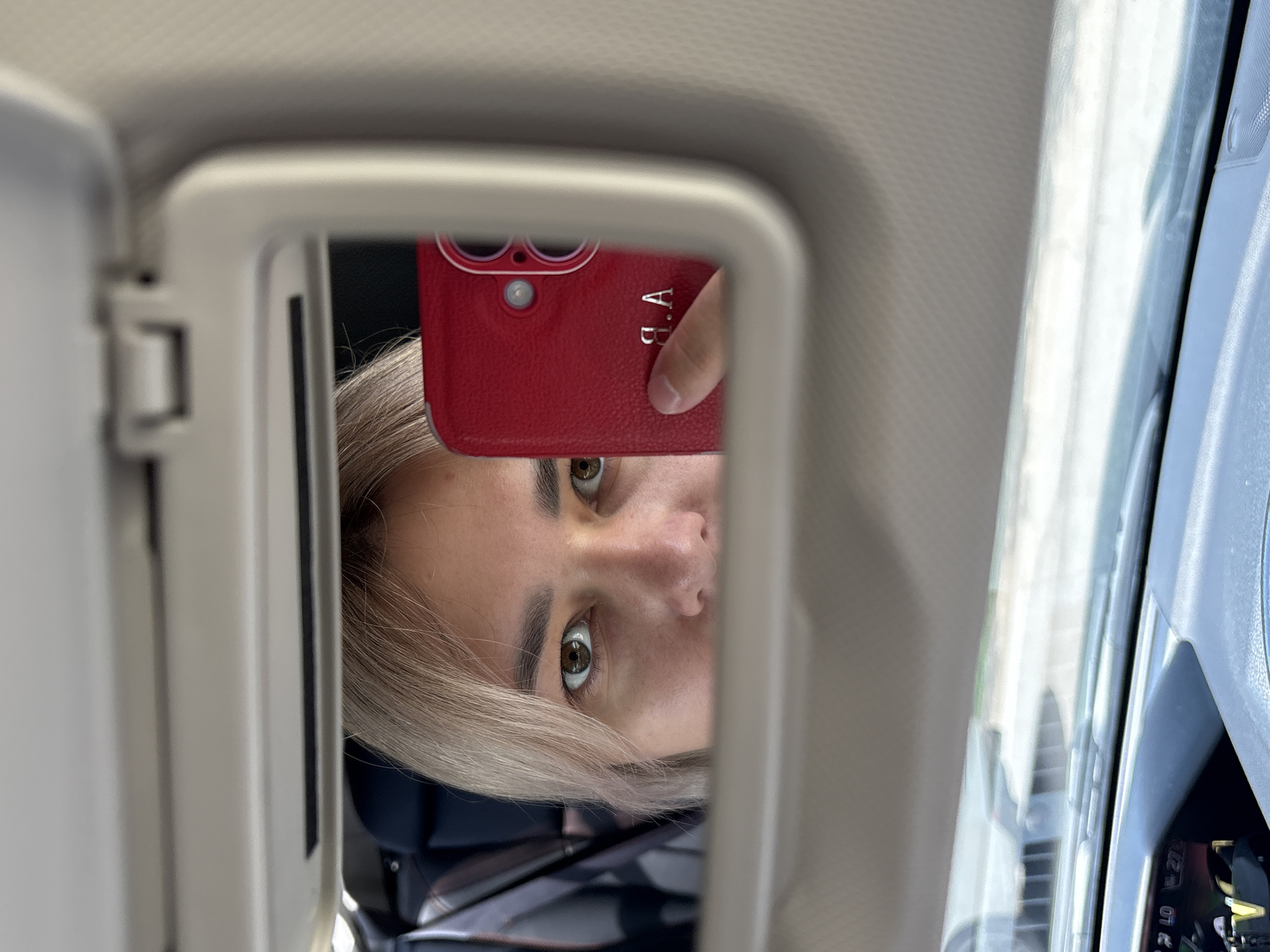 A young woman with blonde hair takes a selfie in a car's visor mirror. She is looking at the camera, holding a red phone in her right hand, which is reflected in the mirror. The phone has a silver monogram that appears to read "A.B." She is wearing makeup, and her eyes are brown. The interior of the car is visible in the reflection, including part of a car seat and the dashboard. The scene is indoors, likely in a vehicle parked within the city of Birobidzhan, Russia.