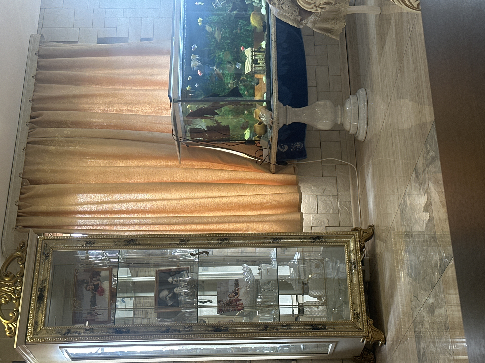 This is a medium shot, eye-level, indoor photograph of a living room. On the left is a tall, ornate, gold-trimmed glass display cabinet filled with glassware and framed photographs. To the right of the cabinet is a large aquarium filled with fish and a miniature Greek-style temple. The aquarium sits on a pedestal that is draped with a blue cloth. In the background are beige curtains and a tiled wall. The floor is tiled with a marble-like pattern. The lighting is natural, coming from the right side of the frame, and casts reflections on the floor. The image depicts a domestic scene, likely in Artemovsky, Russia, given the provided context. There are no people visible in the image.