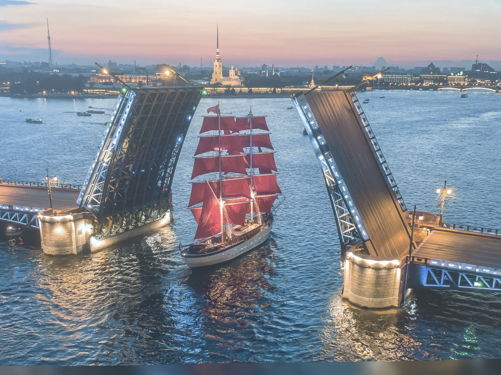 A nighttime scene features a large sailing ship with vibrant red sails passing through a raised drawbridge. The bridge halves are separated to allow the ship to pass. Both sides of the bridge are illuminated with white lights. The ship is positioned in the center of the frame. In the background, a city skyline is visible, including a pointed tower. The water in the foreground and midground reflects the lights of the city and bridge. This scene does not occur in the city of Sochi, Russia. It is most likely the Scarlet Sails celebration in Saint Petersburg, Russia.
