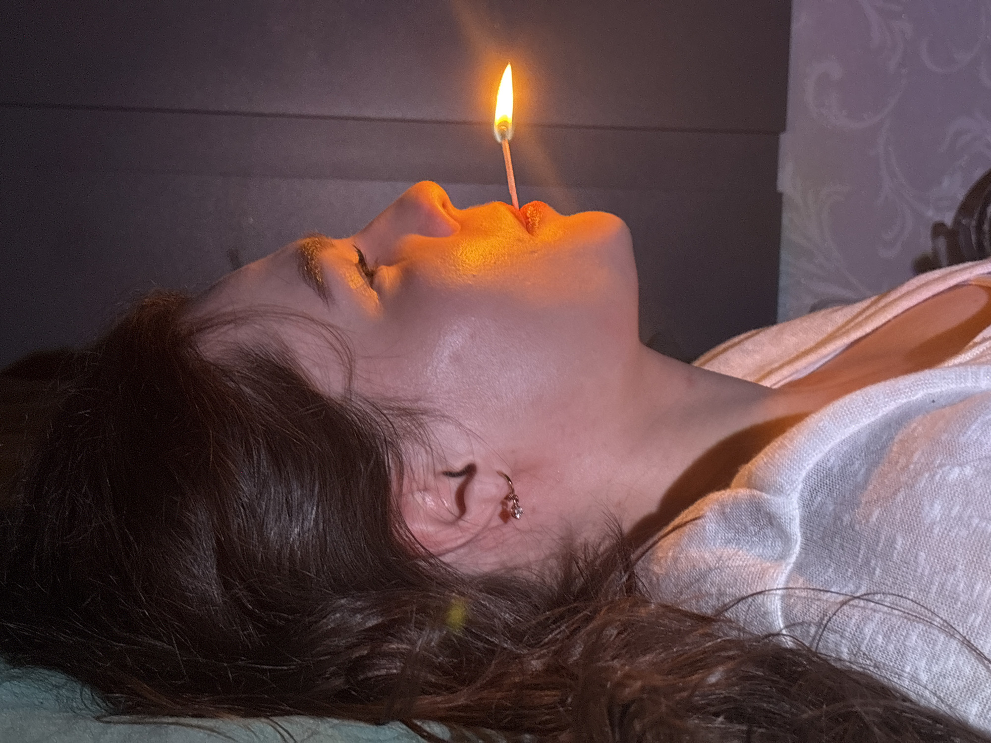 A woman lies on her back, her head tilted slightly upward. A lit matchstick is balanced between her nose and upper lip. The light from the flame illuminates her face. She has dark hair, fair skin, and is wearing a white top. An earring is visible in her left ear. The background includes a dark headboard and patterned wallpaper. The specific location is unknown.