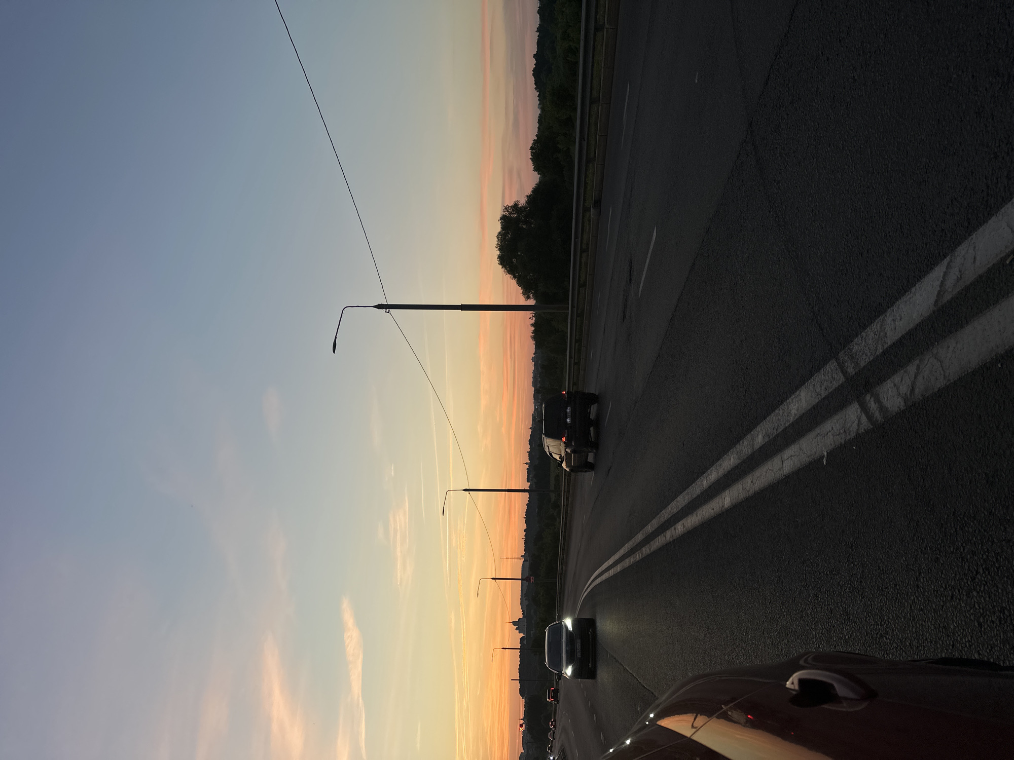 The image depicts a road in Vladimir, Russia, during a sunset. The sky displays a gradient of colors, from soft pinks and oranges near the horizon to a pale blue overhead. The road itself is asphalt, marked with white double lines. Several cars are visible on the road, some in motion and others parked. Streetlights line the side of the road, casting a soft glow. In the background, trees and a few buildings are silhouetted against the twilight sky. The scene suggests a typical evening commute on a highway on the outskirts of the city.