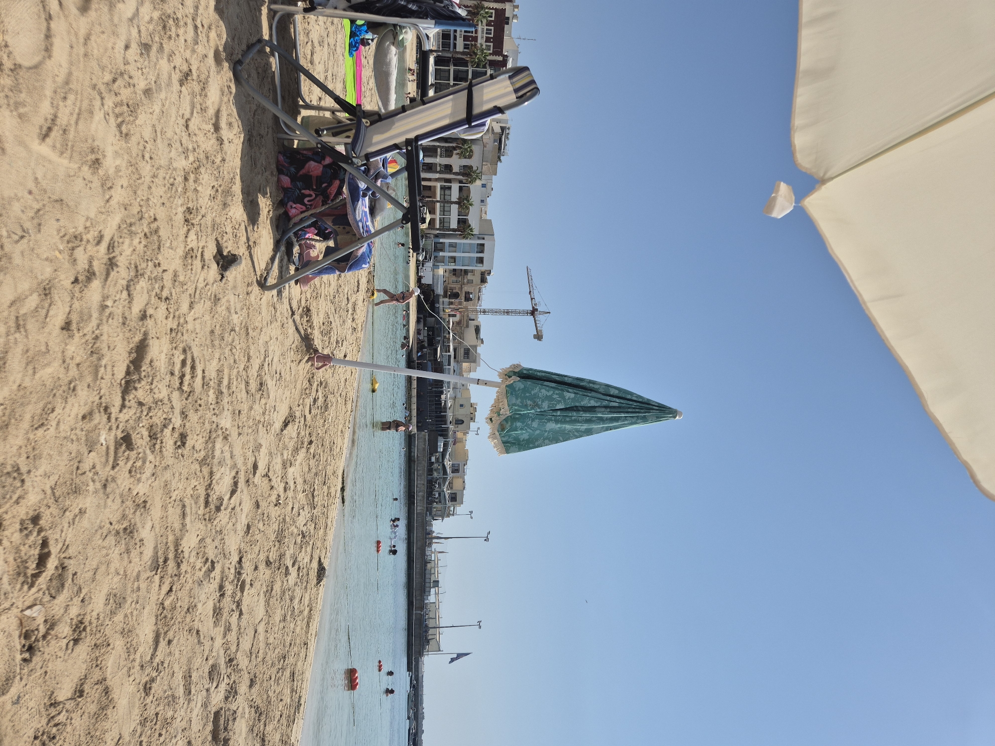 The image depicts a sunny day at a beach in Birżebbuġa, Malta. In the foreground, two beach chairs are set up on the sand, one with a blue and white striped pattern, and the other partially obscured and covered with a beach towel featuring a flamingo pattern. A closed, teal-colored umbrella with white fringe stands to the right of the chairs. Beyond the chairs, the shallow, clear water of the sea stretches out, with several people swimming and enjoying the water. Orange buoys mark a designated swimming area. In the background, a coastal town with multi-story buildings lines the shore, with palm trees visible amongst the architecture. A construction crane is prominently visible in the distance, suggesting ongoing development in the area. The sky is a clear, bright blue.