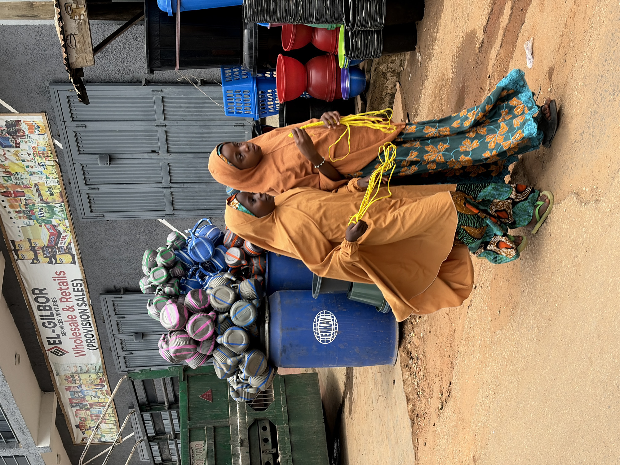 In the city of New Stadium, Nigeria, two young girls, likely siblings, are walking on a dusty, paved street. The girl on the right, wearing an orange hijab and a patterned blue and green dress, holds a yellow rope coiled in her hands. Her younger sister, on the left, also wears an orange hijab and a matching patterned dress, and is carrying a similar yellow rope. They are walking past a blue barrel with a white logo that reads "ENKAY". In the background, a variety of plastic containers, including buckets and bowls in red, blue, green, and black, are stacked. There's also a green truck with a license plate that reads "PLATEAU SA-36XB" and a storefront with a banner that says "EL-GILBOR SERVICES VENTURES WHOLESALE & RETAILS (PROVISION SALES)". The scene is likely a commercial area given the goods displayed and the store signage.
