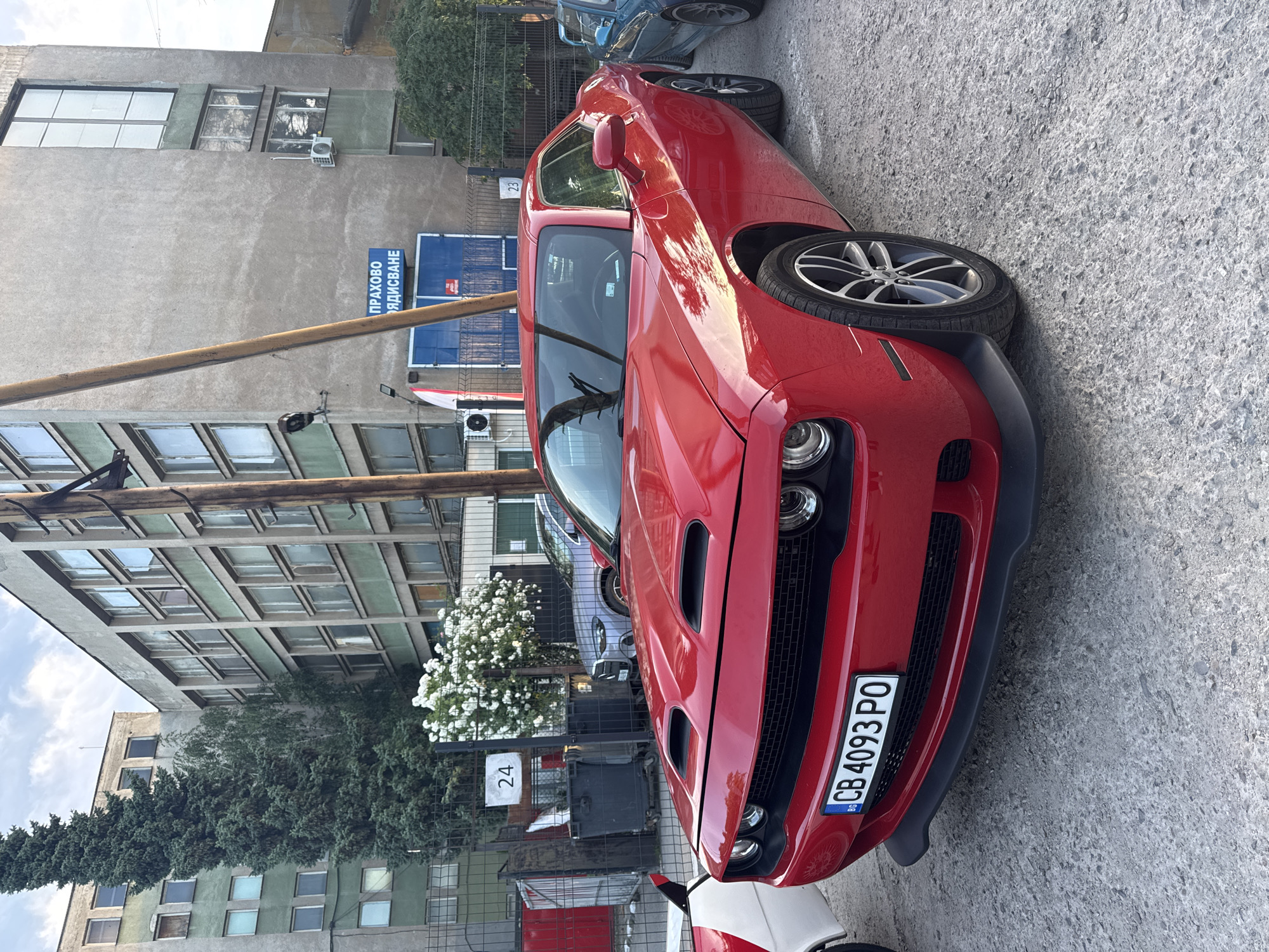 A red Dodge Challenger is parked on a gravel surface in front of an industrial-style building. The car's license plate reads "CB 4093 PO". To the left of the Challenger, a white and red vehicle is partially visible. In the background, the building has numerous windows and a sign with Cyrillic text that reads "ПРАХОВО БДИСКАНЕ". There are two large trash bins to the left of the building, and a tall evergreen tree stands next to the building. A blue car is visible to the right of the Challenger. The scene appears to be in Busmantsi, Bulgaria.