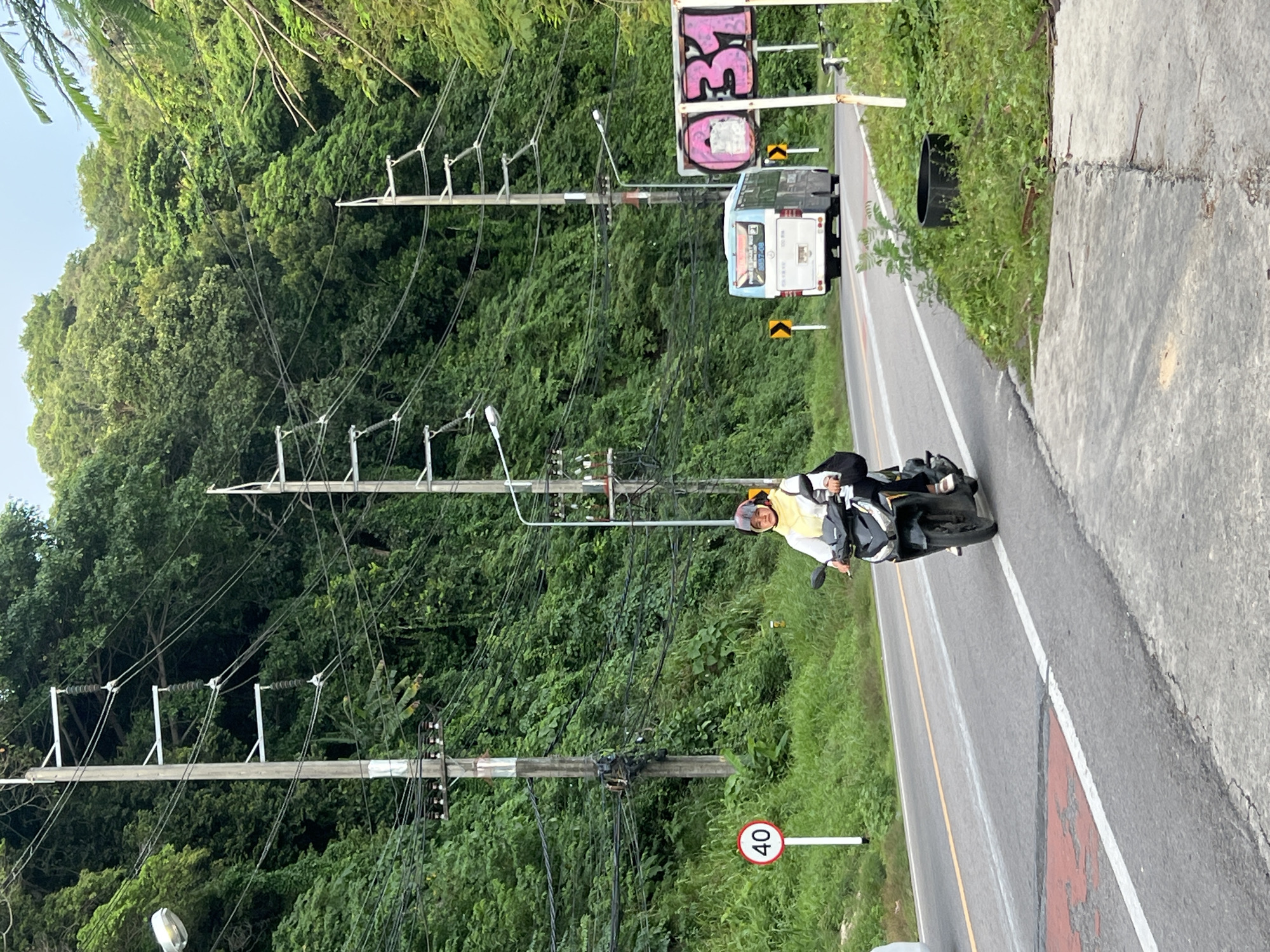 A person rides a black scooter on a paved road in Ban Bang Thao, Thailand, with a green, forested hillside in the background. The person wears a yellow top and a helmet. A white and blue bus is visible further down the road. Road signs, including a speed limit sign indicating "40," are present. Power lines with multiple poles are strung across the scene. The environment is lush with green vegetation.
