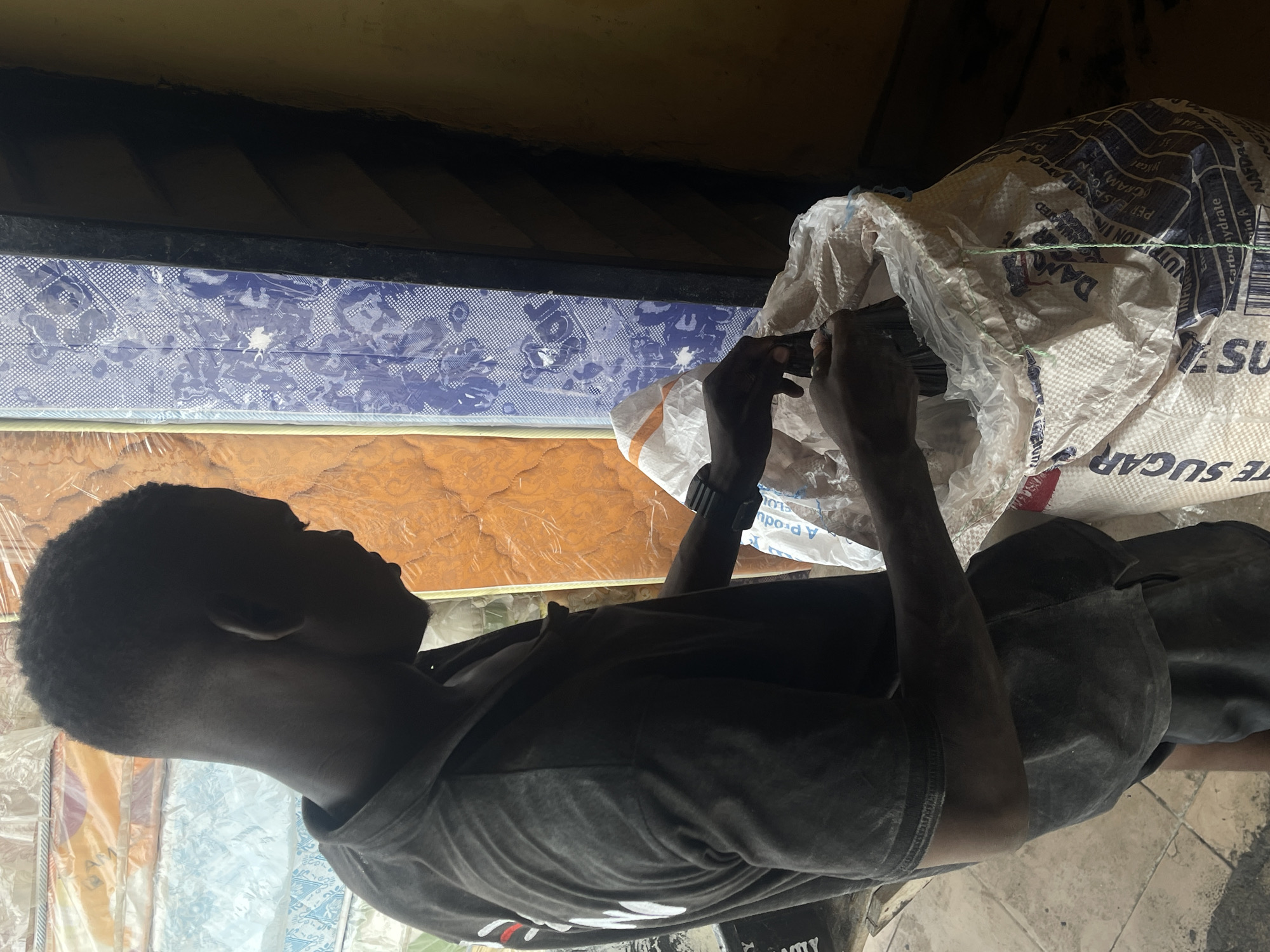 A young Black male is standing in a dimly lit indoor space, possibly a shop or storage area in Ado, Nigeria. He is wearing a dark grey t-shirt and dark shorts. His hair is cut short. He is facing to his right, looking down as his hands are reaching into a large white and orange woven bag. The bag appears to be made of polypropylene and has printing on it, including the words "WHITE SUGAR" and "DANGER" visible. He seems to be handling something within the bag.
Behind him, stacked vertically, are several large, flat items wrapped in plastic. These appear to be mattresses or similar bedding products. The mattresses have patterned covers in blue and white, and orange and yellow. To the right of the stacked items is a dark vertical structure, possibly a doorway or partition, with diagonal yellow and dark brown stripes. The overall lighting is dim, casting shadows and highlighting the textures of the objects. The floor is tiled with a light-colored material.