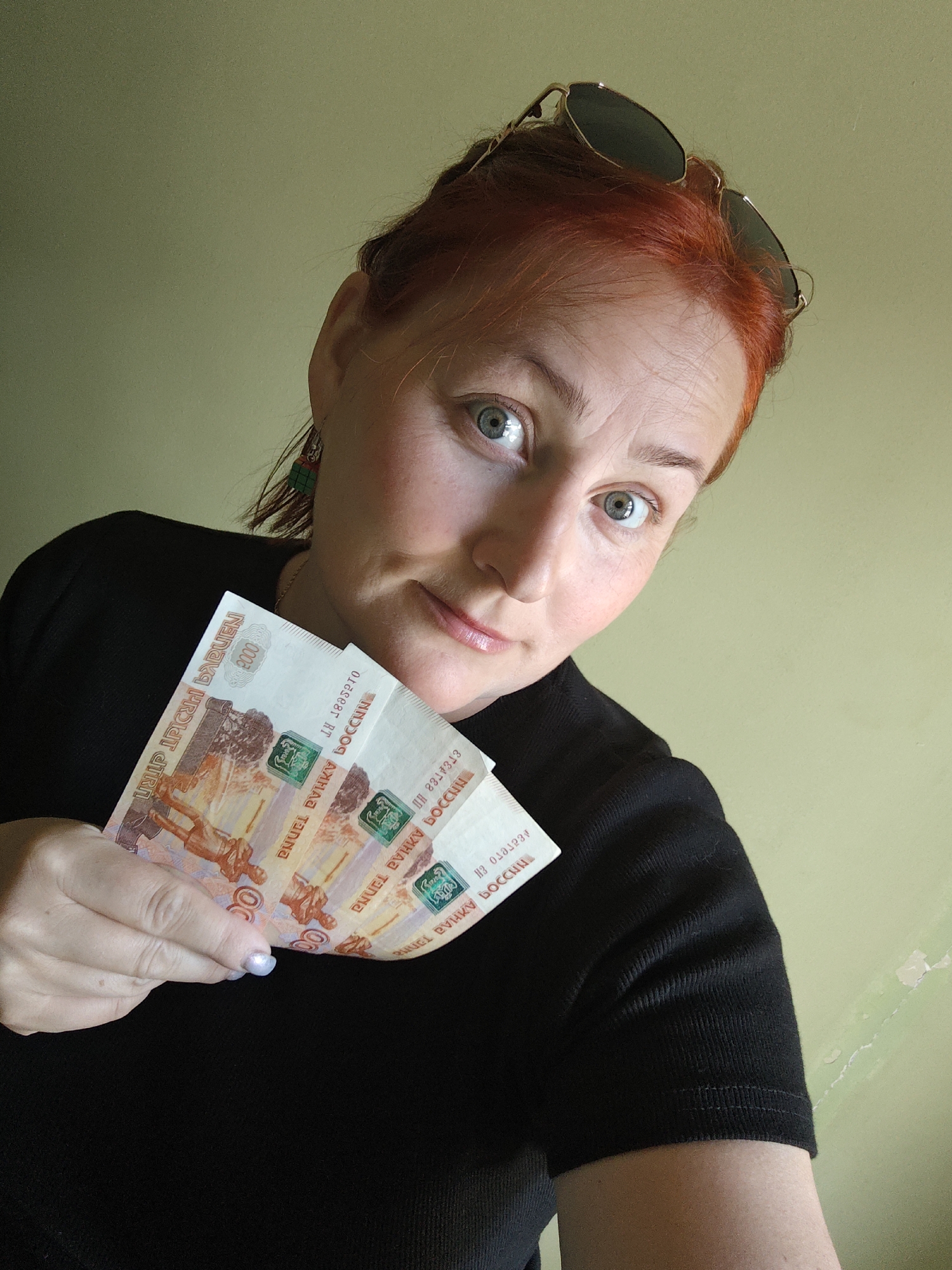 The image shows a woman indoors with light skin and red hair, holding Russian currency (5000 Ruble banknotes) in her left hand. She wears a black top and sunglasses are placed on her head. The background features a light green wall. The OCR of the image is 'ДЕСГего Ен 8784788 ΗΠ PHUCL RVKY OCCHN PNUEL PUHKU BOCCHN BOCCHN AH TERNA OC RT 2000 BARUEN A PRINT STRA'. There is no information about the image being taken in Марковский, Russia.