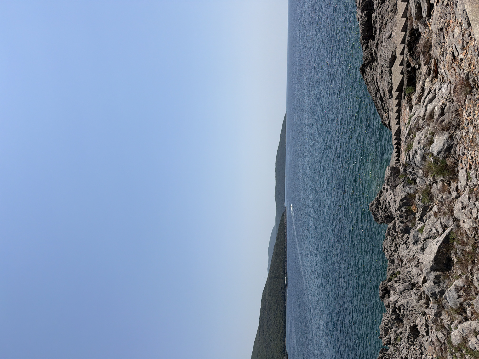 This is a vertical, wide shot of the Bigova bay in Montenegro on a clear, sunny day. The foreground is dominated by a rocky shoreline with a set of concrete stairs leading down to the water on the right. The sea is a deep blue with small waves and sparkling sunlight reflecting off its surface. In the middle ground, a green, forested hill slopes down to the water on the left. A sailboat is anchored in the bay, and a small boat is moving across the water. The horizon is a clear blue sky, with a few distant hills visible on the right.