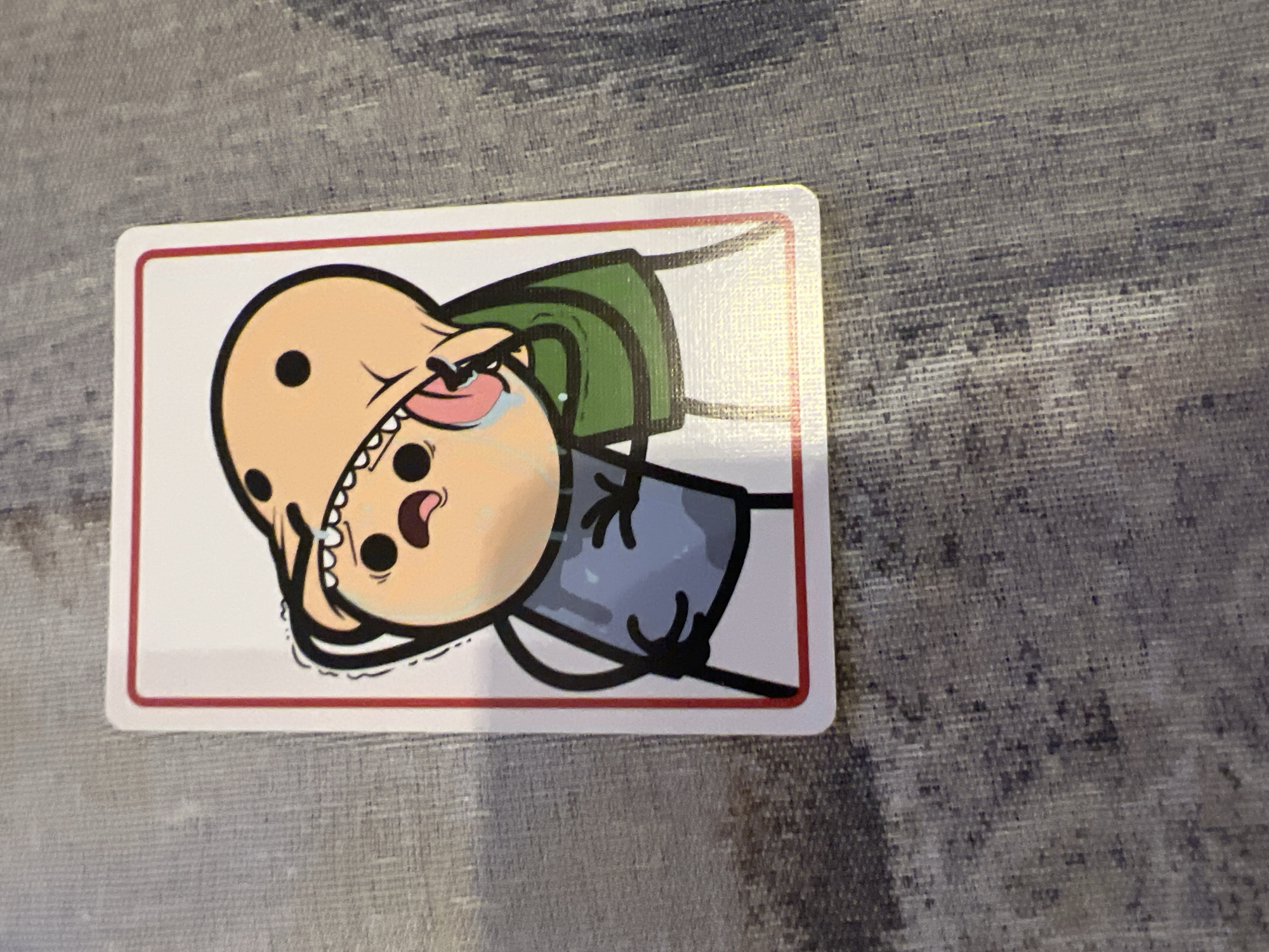 A horizontal, rectangular card with rounded corners is centrally featured, displaying a cartoon character in distress. The card has a distinct red double border on a white background. The character, drawn in a simple yet expressive style with thick black outlines, has light skin, a bald head adorned with several black dots, and small black dot eyes. He is wearing a short-sleeved green top and blue-grey trousers. His mouth is wide open, revealing jagged white teeth and a pink tongue, as if he is screaming or crying out, with a white tear visible near one eye. The character's body is depicted in a somewhat slumped posture.
The card rests on a textured surface, possibly a fabric placemat or tablecloth, exhibiting a mottled pattern of various shades of grey. The lighting is even, suggesting an indoor setting. No discernible text is visible on the card or in the background. The art style is characteristic of webcomics, often used in satirical or dark humor contexts. This image was taken in Plovdiv, Bulgaria.
