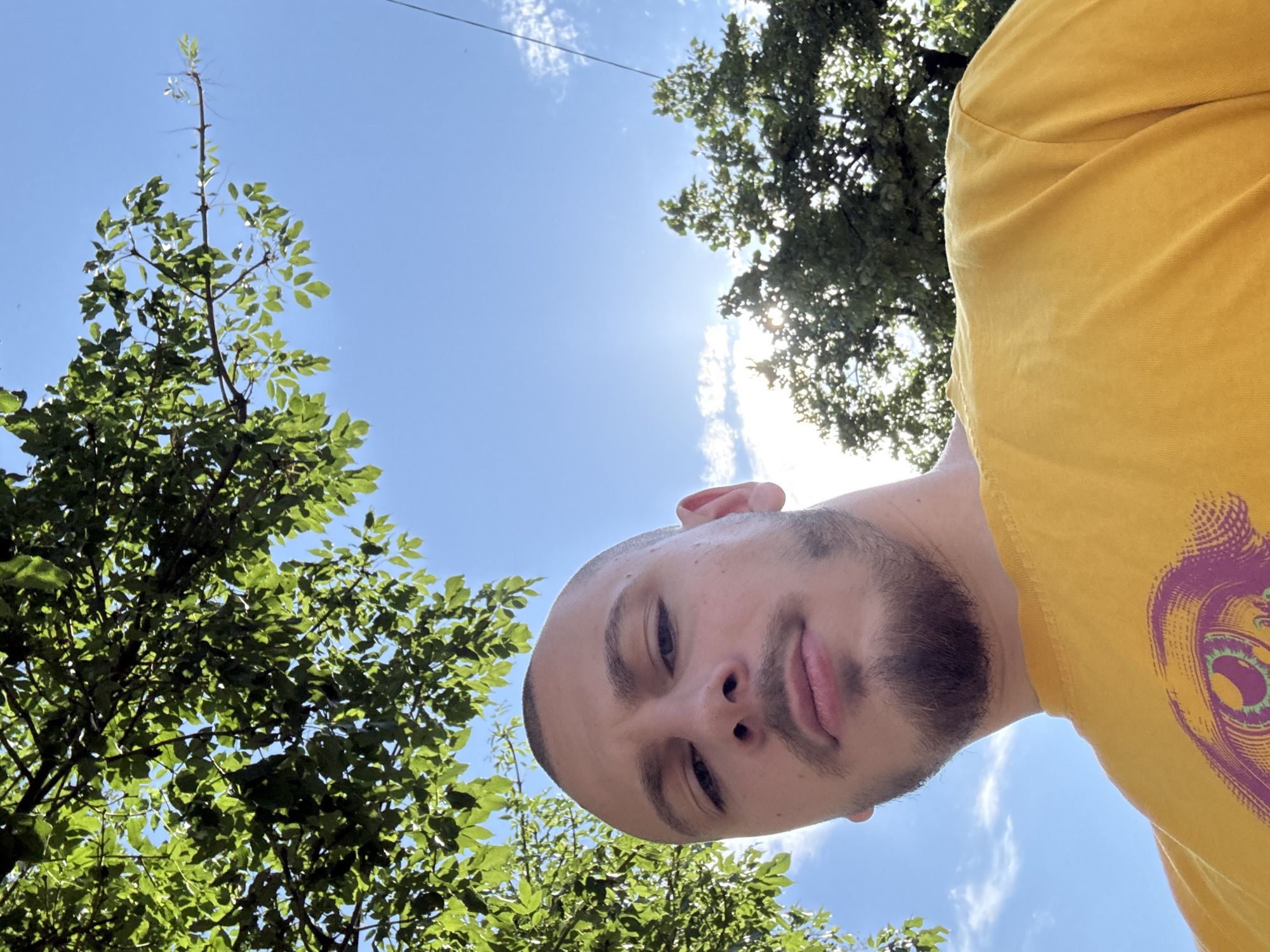 This image is a low-angle, close-up selfie of a male individual in Stara Zagora, Bulgaria, taken outdoors under a bright, partly cloudy blue sky. The subject has a shaved head and a short beard, looking directly at the camera with a neutral expression. He is wearing a plain yellow t-shirt that features a purple and green eye-like graphic design on its lower left. Lush green tree foliage is visible overhead and in the background, with the sun creating a bright glare in the upper right quadrant of the image. A thin dark line, consistent with a utility cable, is visible against the sky in the far upper right.