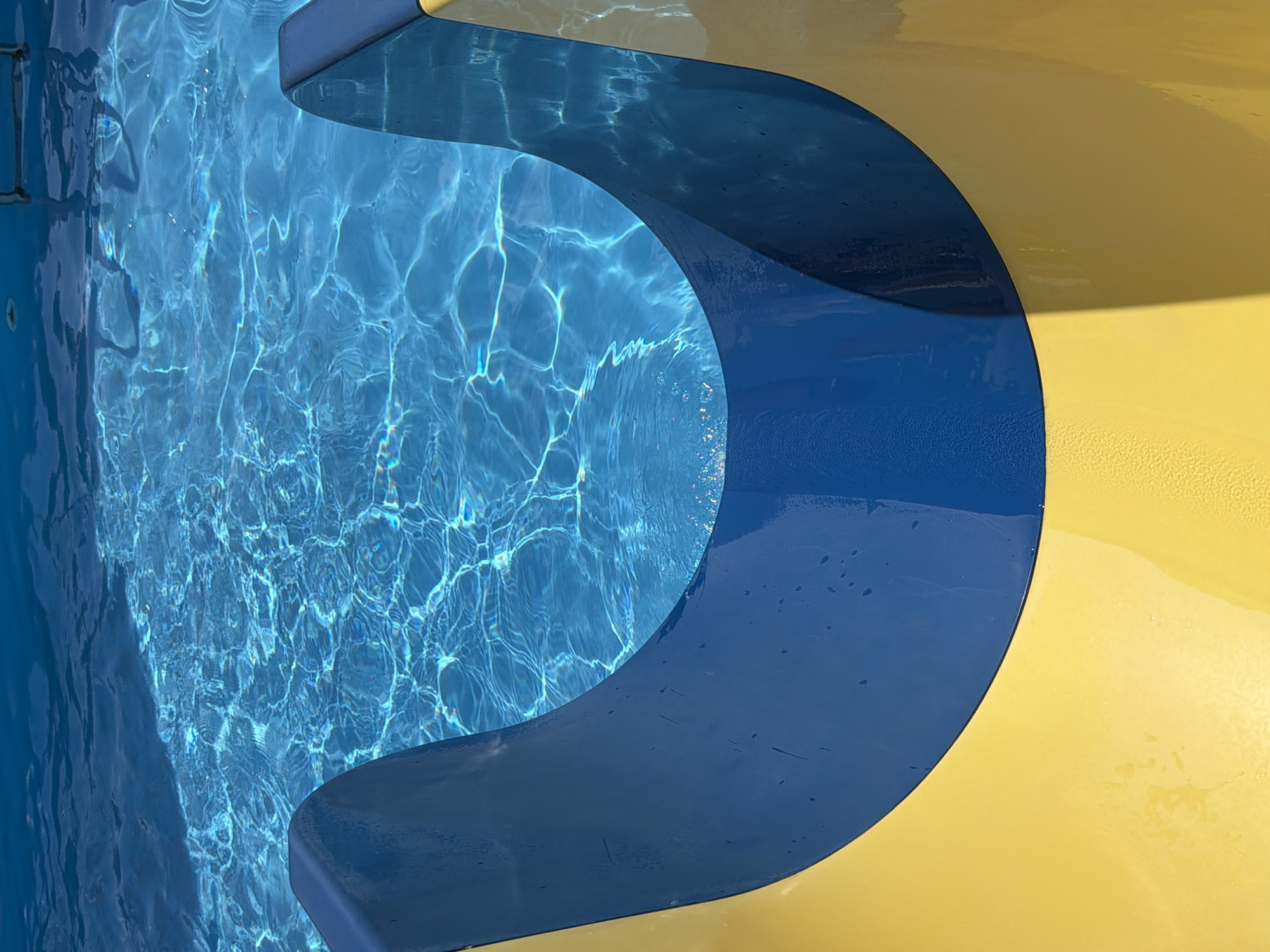 The image shows a close-up view of a section of a bright blue swimming pool with sunlight glinting off the water's surface. Partially visible in the foreground are the curved edges of a yellow and blue water slide, creating a U-shape that frames the pool. The slide's surface appears wet, with small droplets visible. The composition focuses on the textures and colors of the pool water and the slide, suggesting a warm, sunny day at an outdoor recreational facility. The location in Kamyshin, Russia, is not visually evident from the image itself.