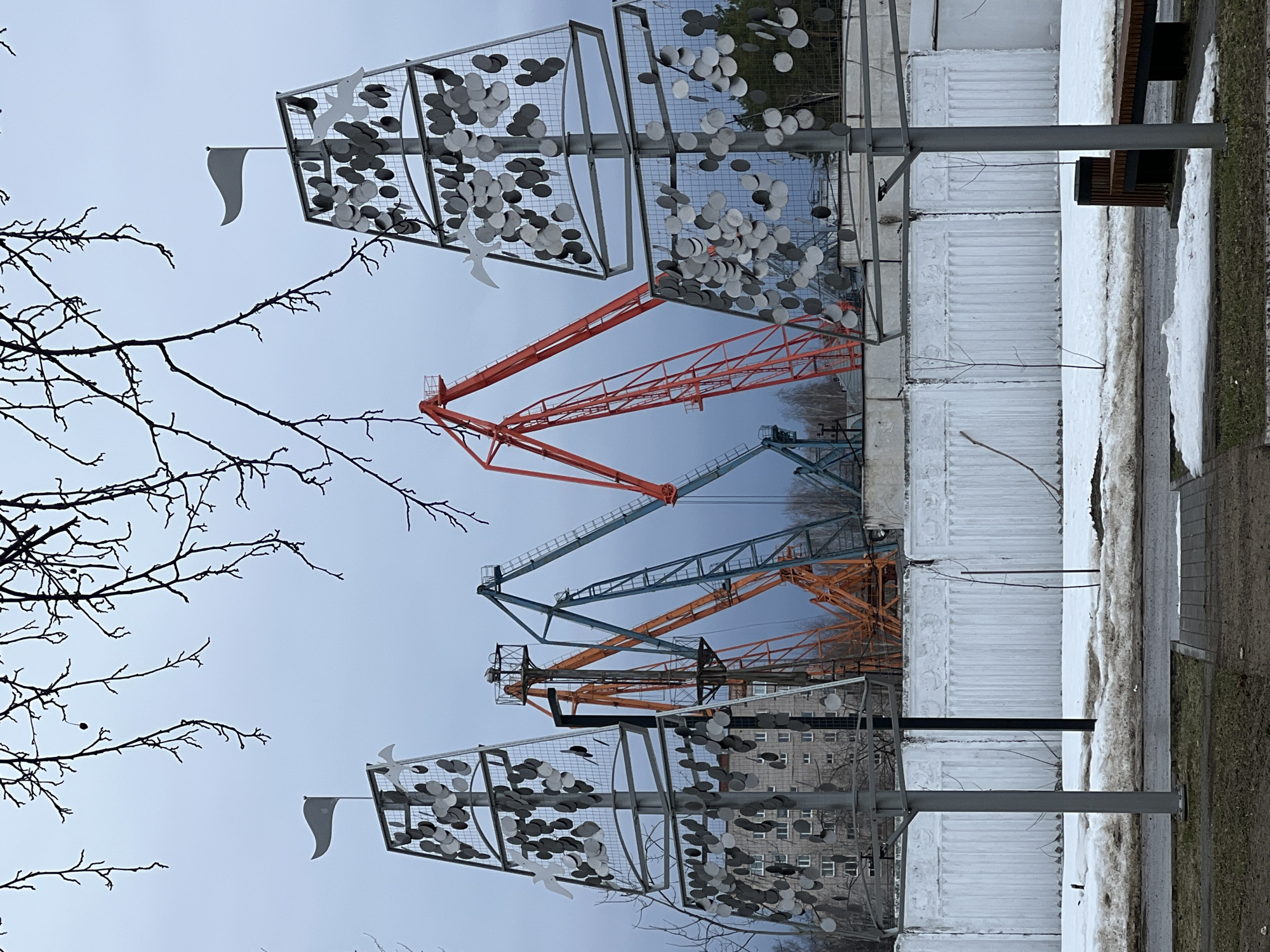 The image depicts an outdoor scene in Chaikovsky, Russia. The sky is overcast and grey. In the foreground, there are metal sculptural installations, potentially public art, that feature wire frames filled with numerous grey discs and white bird shapes. Each sculpture has a small white flag attached. Bare tree branches extend into the frame from the left side.
Behind the sculptures, the outlines of several large construction cranes are visible, with a mix of orange and teal-colored arms. Further back is a multi-story apartment building. In the immediate foreground, there is a concrete barrier or wall with snow accumulated along it. Next to the barrier is a patch of dirt or grass with more snow. A bench with a dark colored seat is affixed near the barrier.