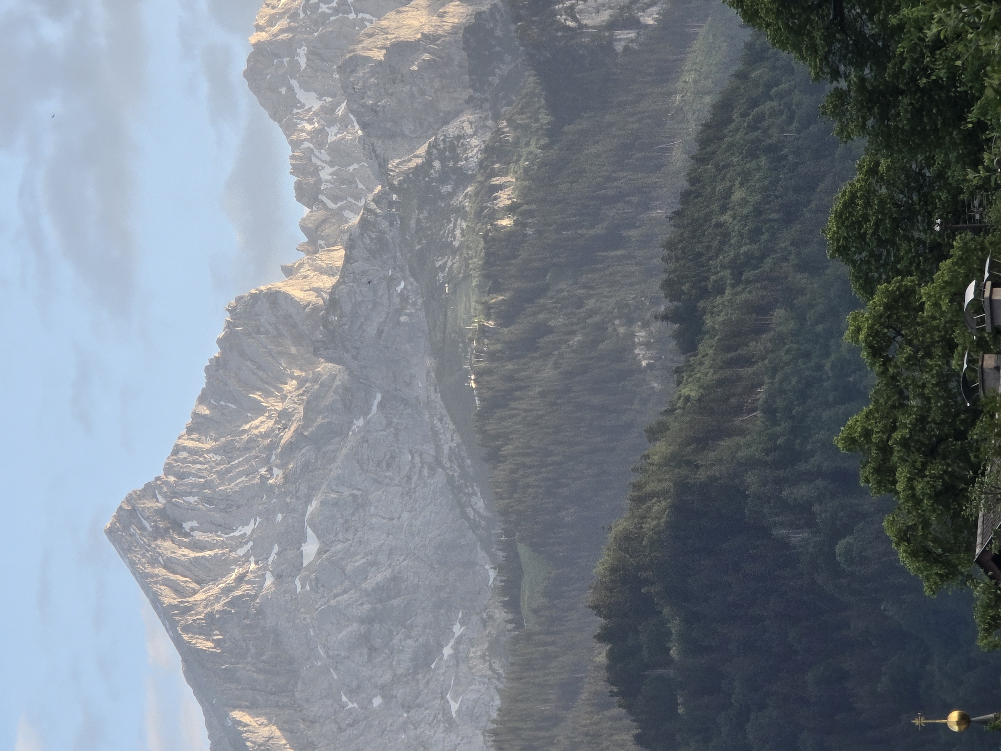 This is a landscape photograph taken in Garmisch-Partenkirchen, Germany. The image depicts a majestic mountain, likely part of the Alps, with rocky slopes and patches of snow. The mountain is bathed in warm, golden sunlight, suggesting it is either early morning or late afternoon. To the right of the mountain, a dense forest of evergreen trees stretches down the slope, fading into a hazy backdrop. Some structures, possibly buildings or a church spire with a golden dome, are visible amidst the trees on the far right, hinting at a settlement or town below. The sky is a pale blue with wispy clouds, and a few birds are seen flying in the distance. The scene evokes a sense of natural grandeur and tranquility. The photograph is oriented vertically, with the mountain occupying the left side and the forest and hint of civilization on the right. There is no visible text in the image.