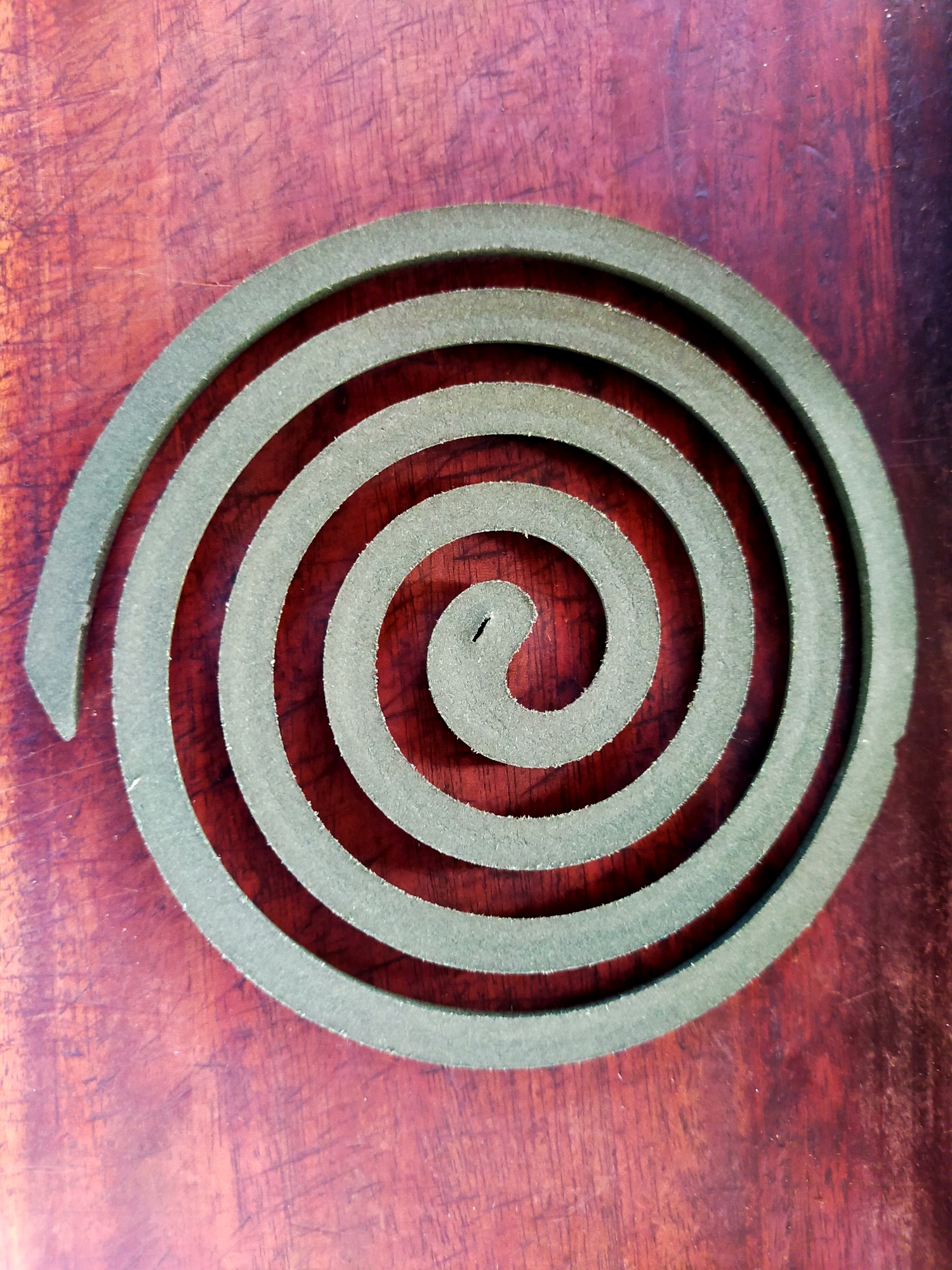 A circular, green mosquito coil lies on a dark, reddish-brown wooden surface. The coil is centrally placed and viewed from directly above. The wood surface exhibits visible grain patterns and some lighter colored scratches or wear marks. The mosquito coil itself is a spiral shape with several concentric turns, made of a powdery, matte material. The lighting is uniform and casts no harsh shadows, suggesting an indoor environment. The image is sharp and clear, focusing on the texture and details of both the coil and the wooden surface. There are no people, animals, or other objects present. Based on the visual information, the location cannot be determined to be Surabaya, Indonesia.