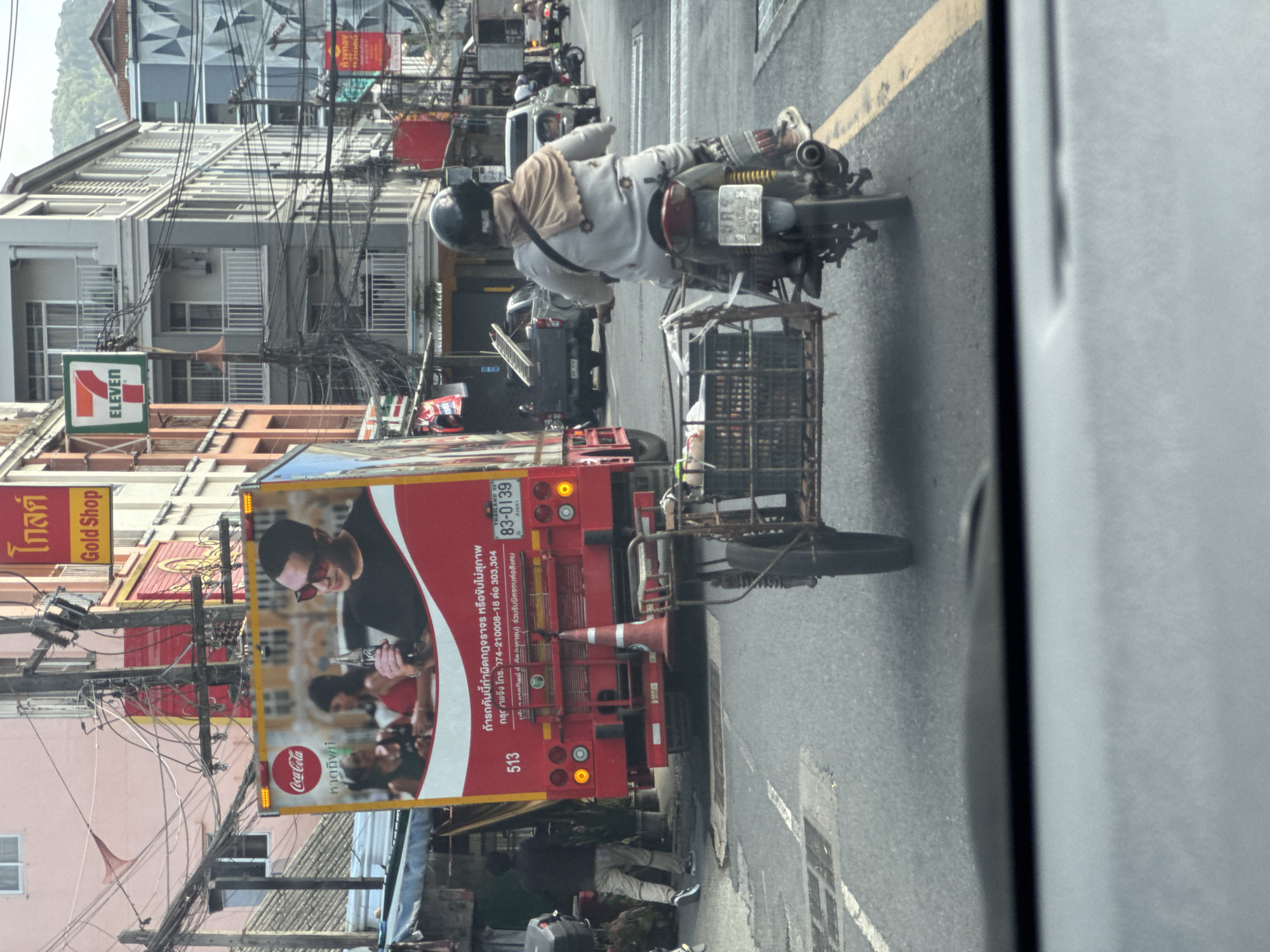 The image depicts a street scene in Patong, Thailand, as viewed from inside a vehicle. Dominating the left side of the frame is the rear of a red Coca-Cola branded truck, featuring an advertisement with individuals holding Coca-Cola bottles. The truck has the license plate "83-0139 THAILAND". To the right of the truck, a person wearing a helmet and a light-colored headscarf and tunic rides a motorcycle with a sidecar containing crates. The motorcycle's license plate is partially visible. In the background, there are multi-story buildings with visible signs, including a "7 ELEVEN" sign and other businesses. Power lines and streetlights are prominent throughout the scene. The road surface appears to be asphalt with visible white and yellow lane markings. The overall atmosphere suggests a busy urban environment.