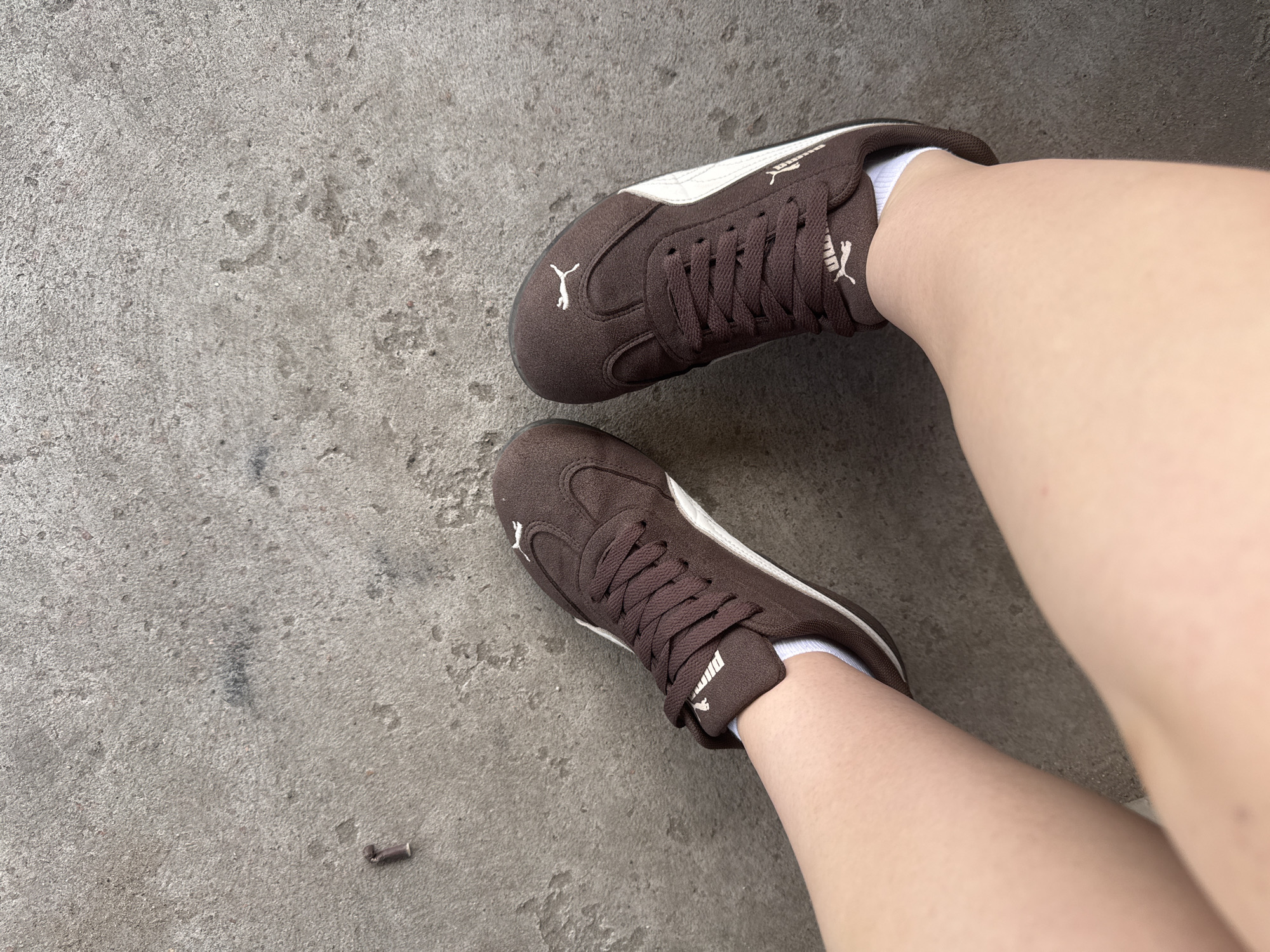 An overhead, medium shot shows a person's legs and feet wearing brown Puma sneakers with white accents and white socks, resting on a concrete surface. The concrete appears weathered and uneven, with a small, dark, cylindrical object lying on it to the left of the frame. The shot is taken from a high angle, looking down at the subject's legs and feet. The location is stated to be in Shulhynka, Ukraine.