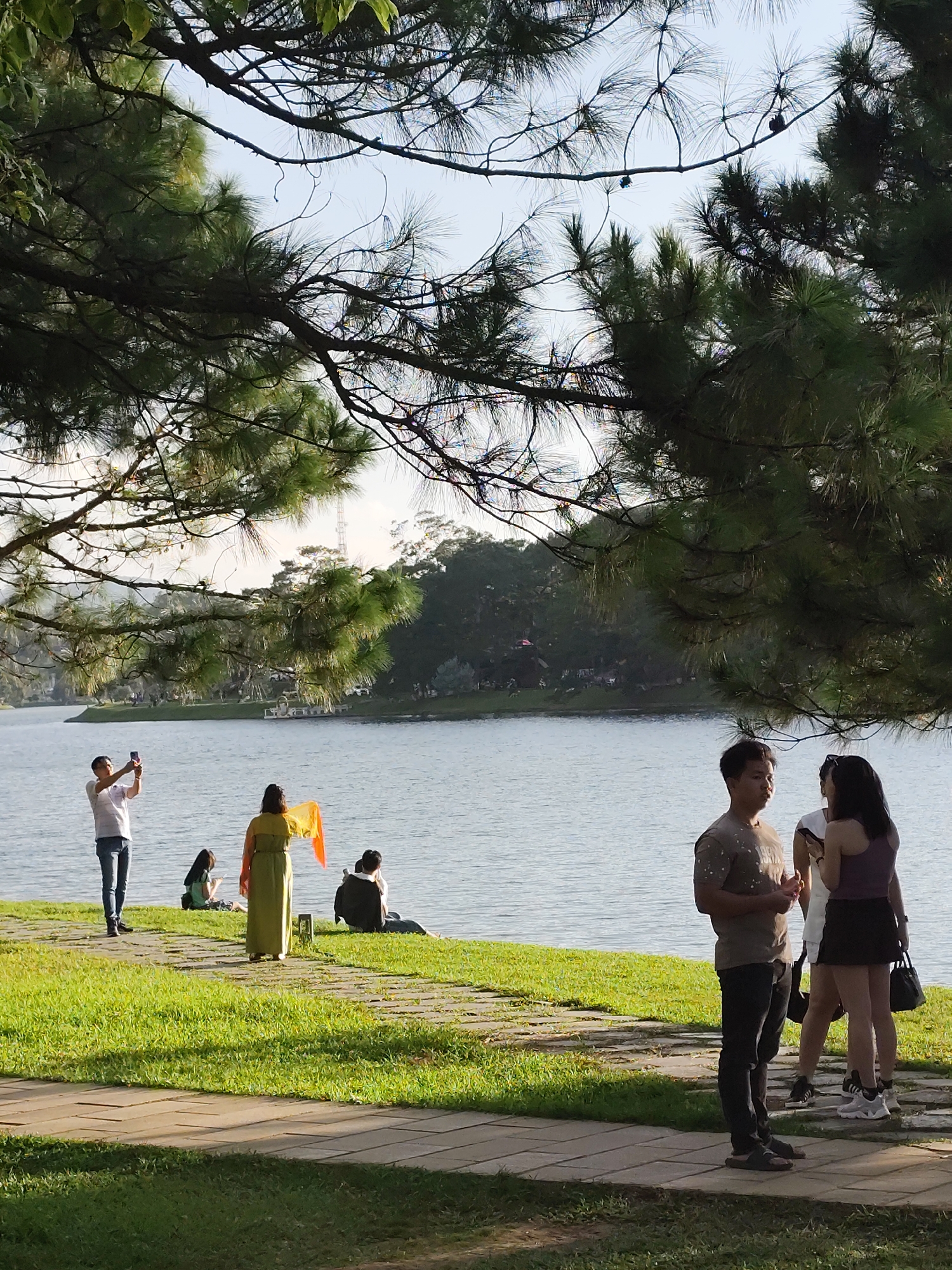 The image depicts a serene outdoor scene at Xuan Huong Lake in Da Lat, Vietnam, captured on a bright, clear day. The foreground is dominated by lush green grass and a paved pathway, partially shaded by the branches of a large pine tree. Several people are scattered along the lake's edge. On the left, a man in a white shirt and jeans stands, holding up his phone as if taking a picture. Nearby, a woman in a long, flowing olive-green dress holds an orange scarf or fabric aloft. Further along the shore, two individuals are seated, one appearing to read or use a device. On the right side of the image, a man in a t-shirt and dark pants stands facing a woman in a purple tank top and dark skirt, engaged in conversation. Another woman in a white top and dark skirt is partially visible behind them. The calm surface of Xuan Huong Lake stretches across the middle ground, reflecting the sky and the distant tree-covered hills. The overall atmosphere suggests a relaxed and leisurely gathering by the water.