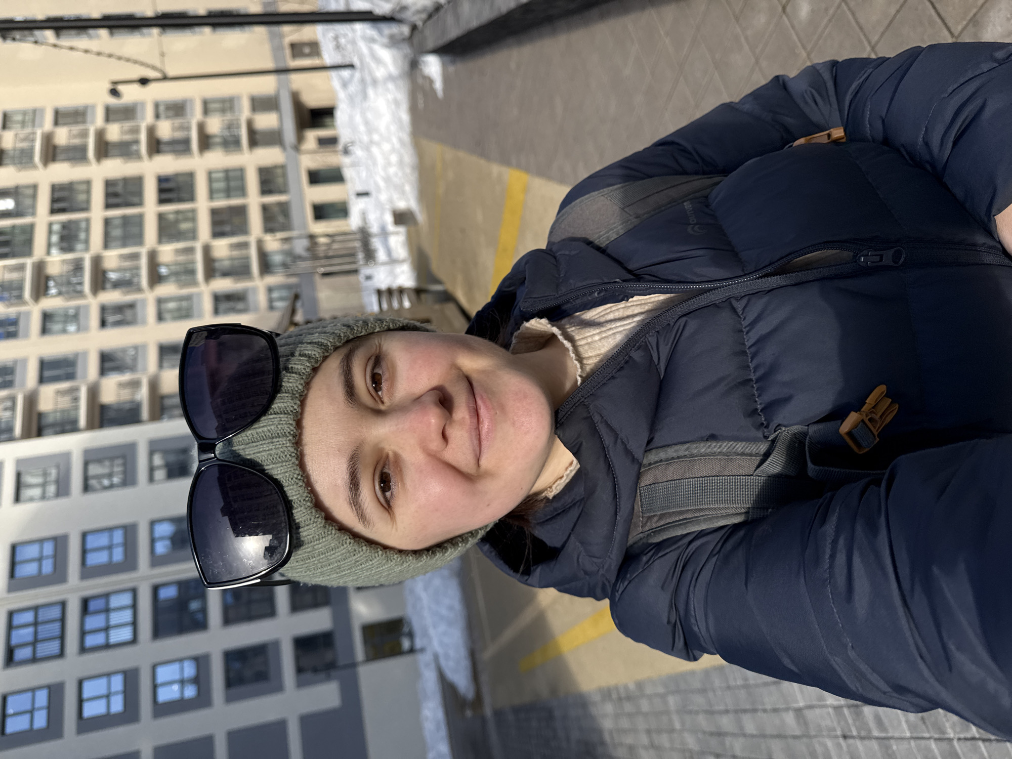 The image depicts a frontal, eye-level outdoor photograph of a female individual within a residential area of Izhevsk, Russia. The subject, a young adult with fair skin and brown eyes, is positioned centrally and exhibits a slight smile. She is wearing a light green knitted beanie, upon which dark-lensed, black-framed sunglasses are perched. Her attire includes a dark blue puffer jacket, partially unzipped, revealing a light-colored, ribbed garment underneath. A grey backpack with brown strap buckles is visible on her right shoulder.
The background features two multi-story apartment buildings. The structure on the viewer's left has a light grey facade with numerous dark-framed windows. The building on the viewer's right, positioned further back, is beige with multiple balconies. The ground in the foreground is paved with light-colored blocks, displaying a prominent yellow line. Patches of snow are present on the ground, particularly to the right of the paved area, indicating cold weather conditions. The scene is brightly illuminated by direct sunlight, casting distinct shadows. A street lamp post is visible in the far right background.
