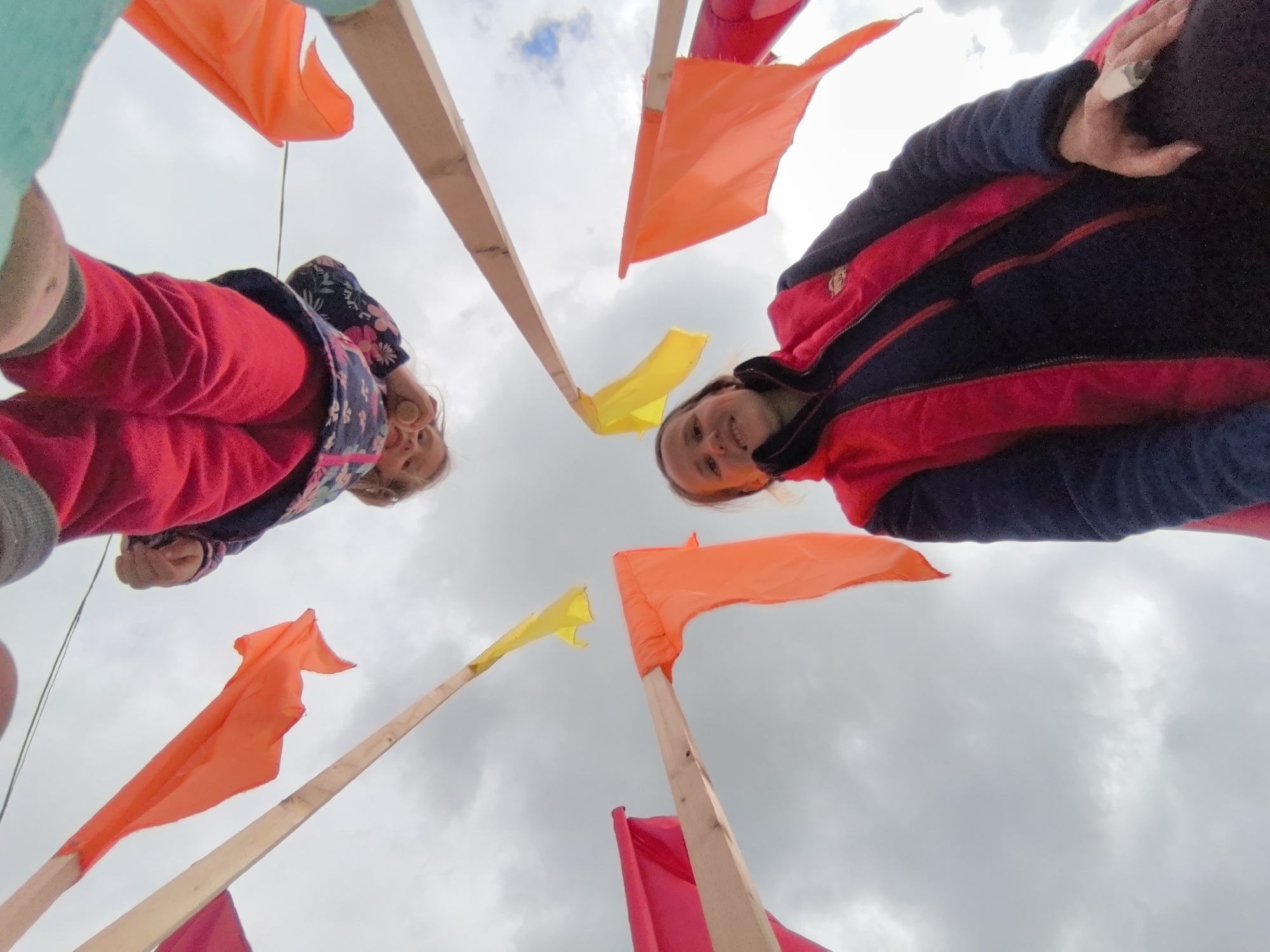 Two individuals, a child and an adult, are looking down towards the camera from above, interspersed with a series of small colorful flags on wooden poles. The sky is visible in the background and is overcast. The location is Shabury, Russia.