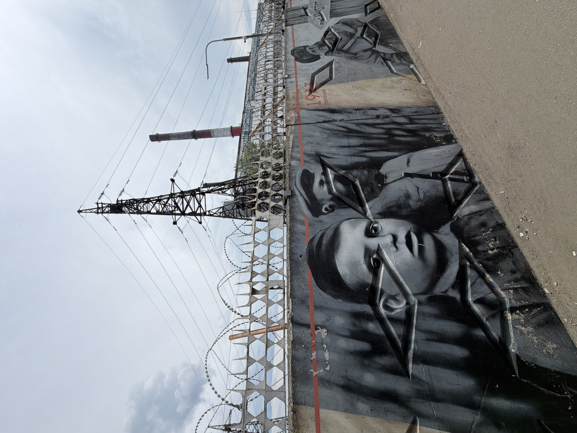 The image depicts a concrete wall with graffiti art in Birobidzhan, Russia. The graffiti features a mural of children's faces in a monochromatic style. In the background, there is a barbed wire fence and industrial structures, including electrical towers and smokestacks, suggesting an industrial or semi-industrial area. The sky is overcast. The wall itself has a red horizontal line running across it. The foreground shows a dirt or gravel path.