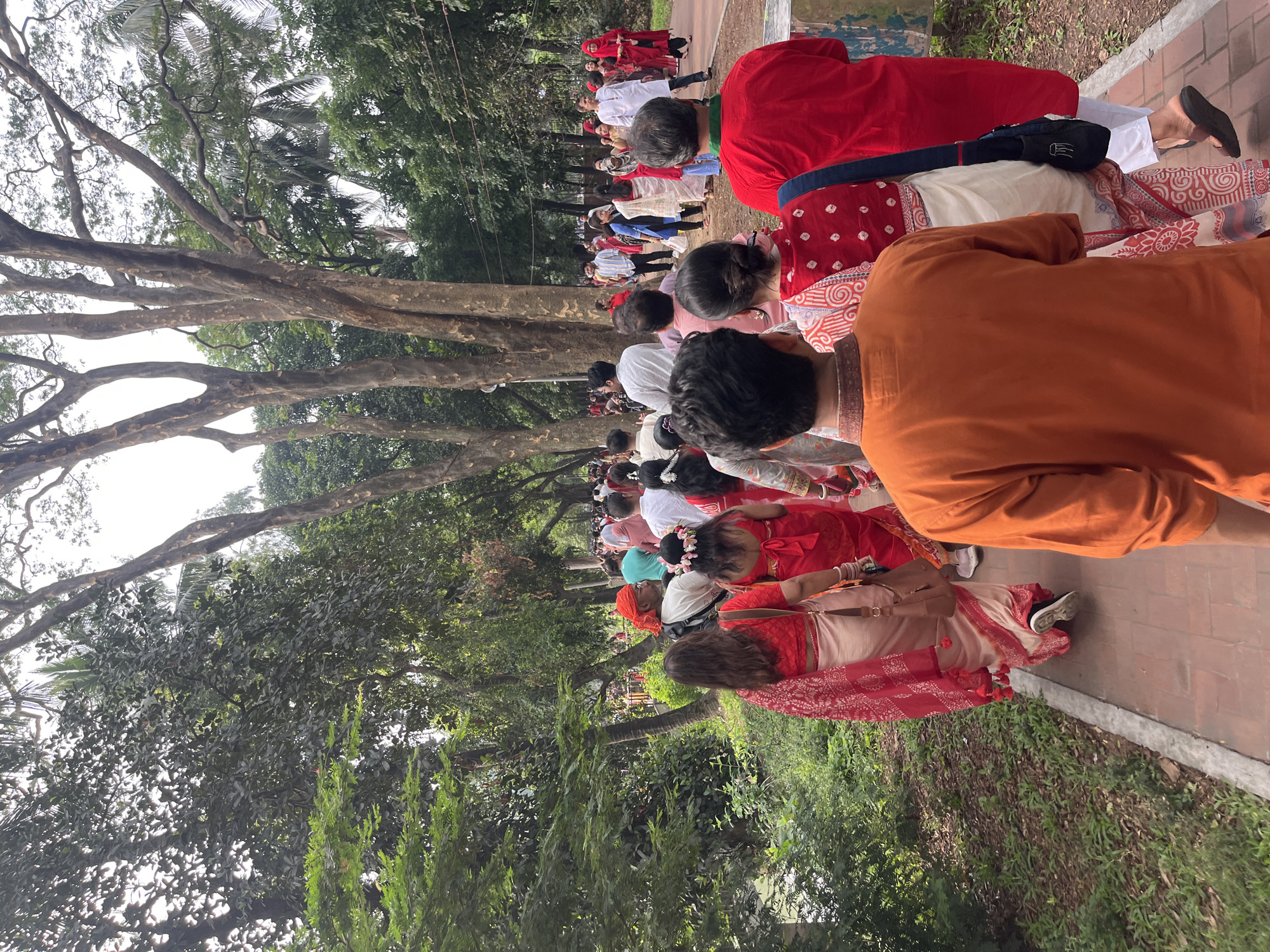 A procession of people walk on a brick-laid path in what appears to be a park setting in Dhaka, Bangladesh. The participants are dressed in traditional clothing, predominantly shades of red, orange, and white, suggesting a cultural event. The path is bordered by lush greenery and tall trees, creating a natural canopy above the crowd. The composition of the image includes individuals in various states of motion, some walking directly towards the camera, others slightly angled away, thus providing a sense of depth and movement. The perspective suggests that the photograph was taken at eye-level from the side of the path, capturing a moment within the flow of the ongoing procession.