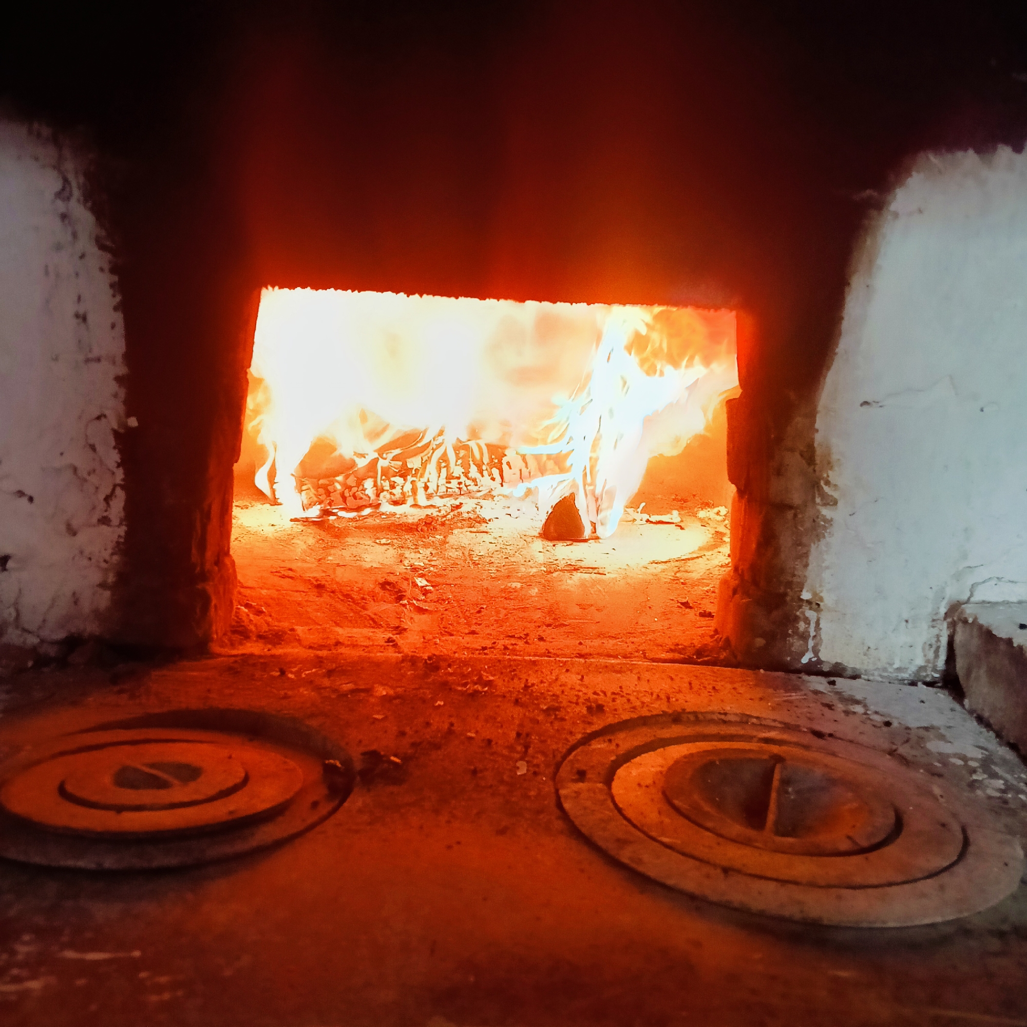 The image depicts an open, brick wood-burning stove with a fire blazing inside. Two round metal plates with concentric rings are visible on the surface in front of the firebox. The surrounding walls are painted white and appear to be aging. There is no indication of any people present. The location, Буренка, Russia, is not explicitly indicated in the image, and is based on user instructions.