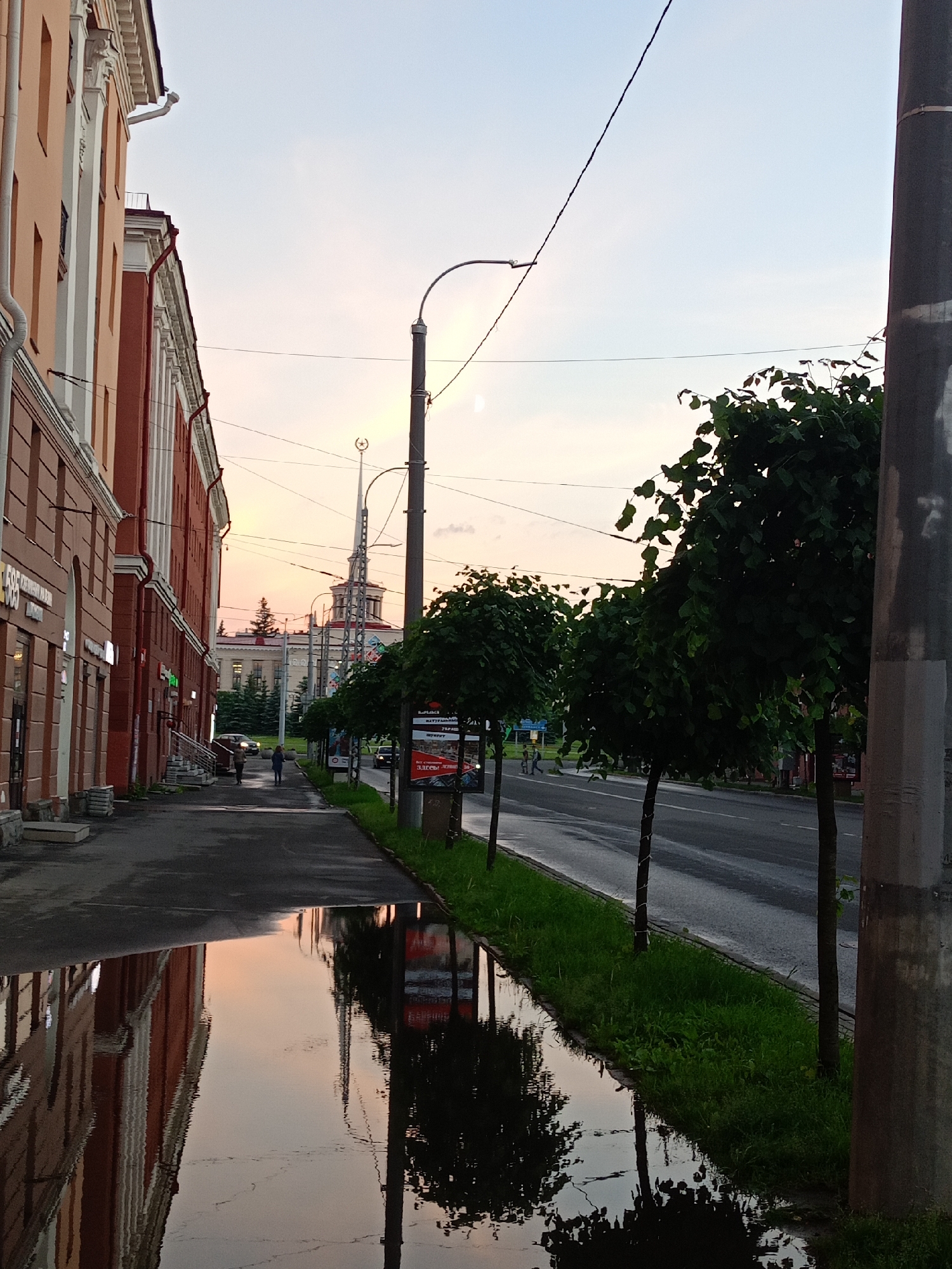 The image depicts a street scene at dusk in a city. On the left, a multi-story building with an orange facade and white trim runs along the sidewalk. The sidewalk is wet and reflects the building and the sky. Several shops with signs are visible at the base of the building. A few people are walking on the sidewalk. On the right side of the street, there are several small, well-maintained trees lining the road. The road itself is wet, suggesting recent rain. In the distance, a tall, ornate tower with a star on top is visible, illuminated by the fading light. The sky is a soft blend of pale blue and pink, indicating either sunrise or sunset. A large, dark utility pole stands prominently in the foreground on the right side of the image. The overall impression is one of a quiet urban street after a rain shower.
