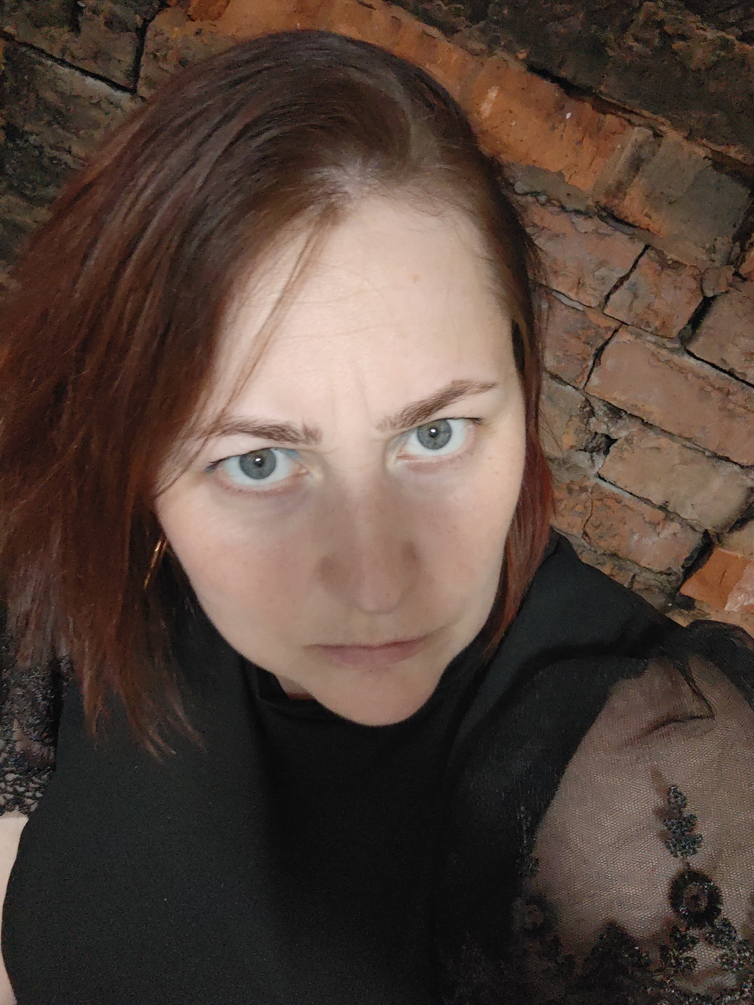 A medium, close-up shot depicts a Caucasian woman with light skin and auburn hair. She looks directly at the camera with a serious expression. Her eyes are light blue, and she is wearing a black top with lace detailing on one of the sleeves.
Behind her is a brick wall.
The location of Фоки, Russia cannot be confirmed from visual data in the image.