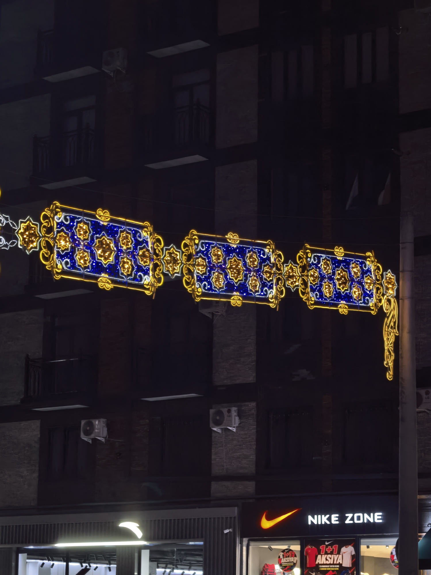 The image depicts a nighttime street scene in Fergana City, Uzbekistan, featuring festive decorative lights strung across a building facade. The lights form a long, illuminated banner composed of rectangular segments, each filled with a blue and gold star pattern. These segments are outlined in a bright yellow, with additional ornate yellow scrollwork embellishing the edges. The building behind the lights appears to be a multi-story structure with dark windows and balconies. Below the banner, a store with a prominent "NIKE ZONE" sign is visible, illuminated by its own neon lights and featuring displays of athletic apparel in its windows. The overall impression is one of an urban commercial area decorated for a special occasion or holiday.