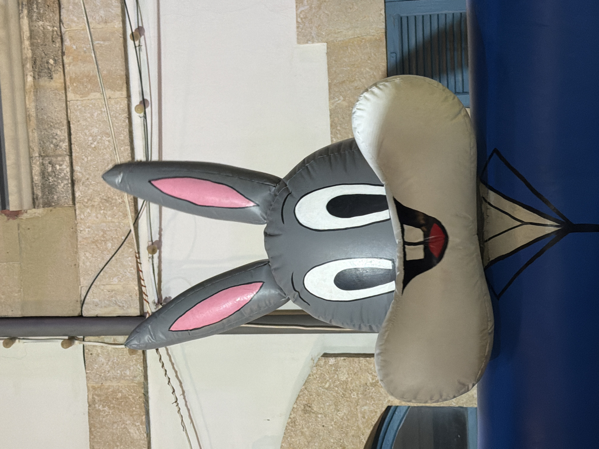 An eye-level, medium shot depicts a gray, inflatable Bugs Bunny head attached to a large blue cylinder in Larnaca, Cyprus. The background features tan and white stucco walls. The Bugs Bunny head has two long gray ears with pink insides, large white eyes with black pupils, and large white cheeks. The bunny head is attached to a blue cylinder with black and white line decorations. Wires and cables run along the wall behind the inflatable.