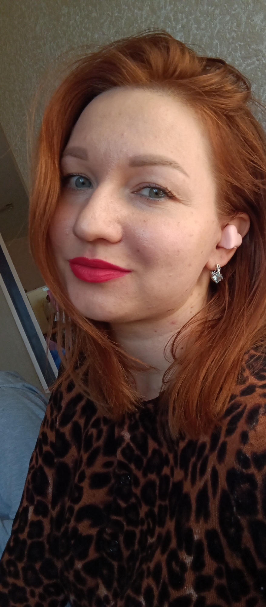 A close-up portrait depicts a woman with red hair, fair skin, and blue eyes. She is wearing red lipstick and a leopard print top. She has a pink heart-shaped object in her left ear and a silver earring on the earlobe. The setting appears to be indoors, with a glimpse of what could be a window or wall in the blurred background. Location is Unknown, Unknown.