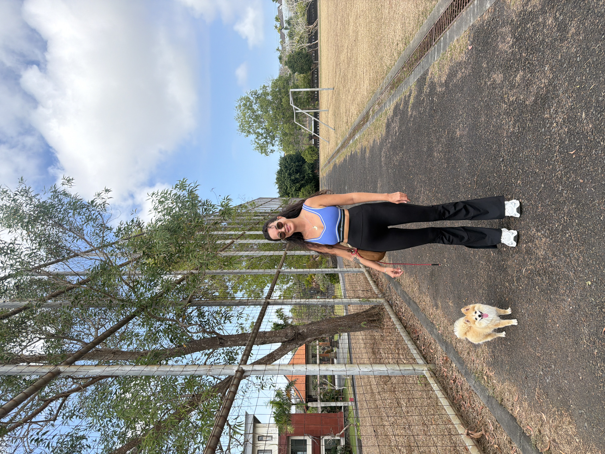In an outdoor scene in Unknown, Unknown, a woman with long, dark, wavy hair and sunglasses is walking a small, fluffy, light brown dog on a leash. The woman is wearing a blue sports bra with white trim and black flare-leg athletic pants, along with white sneakers. She has a brown purse slung over her shoulder. The dog is looking towards the camera with its tongue out.
They are walking on a paved path that runs through an open field. To the left of the path is a chain-link fence with metal posts and trees growing behind it. In the background to the left, there are buildings with red and white facades and palm trees. To the right of the path, the field is covered in dry, yellow grass and has a soccer goal in the distance. The sky is blue with scattered white clouds. The overall lighting suggests a daytime scene.